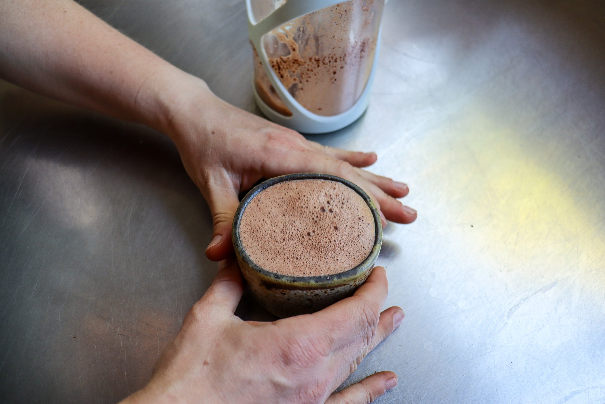 Person holding a ceramic cup filled with artisan drinking chocolate on a metallic surface with a glass pitcher containing remaining drinking chocolate in the background.