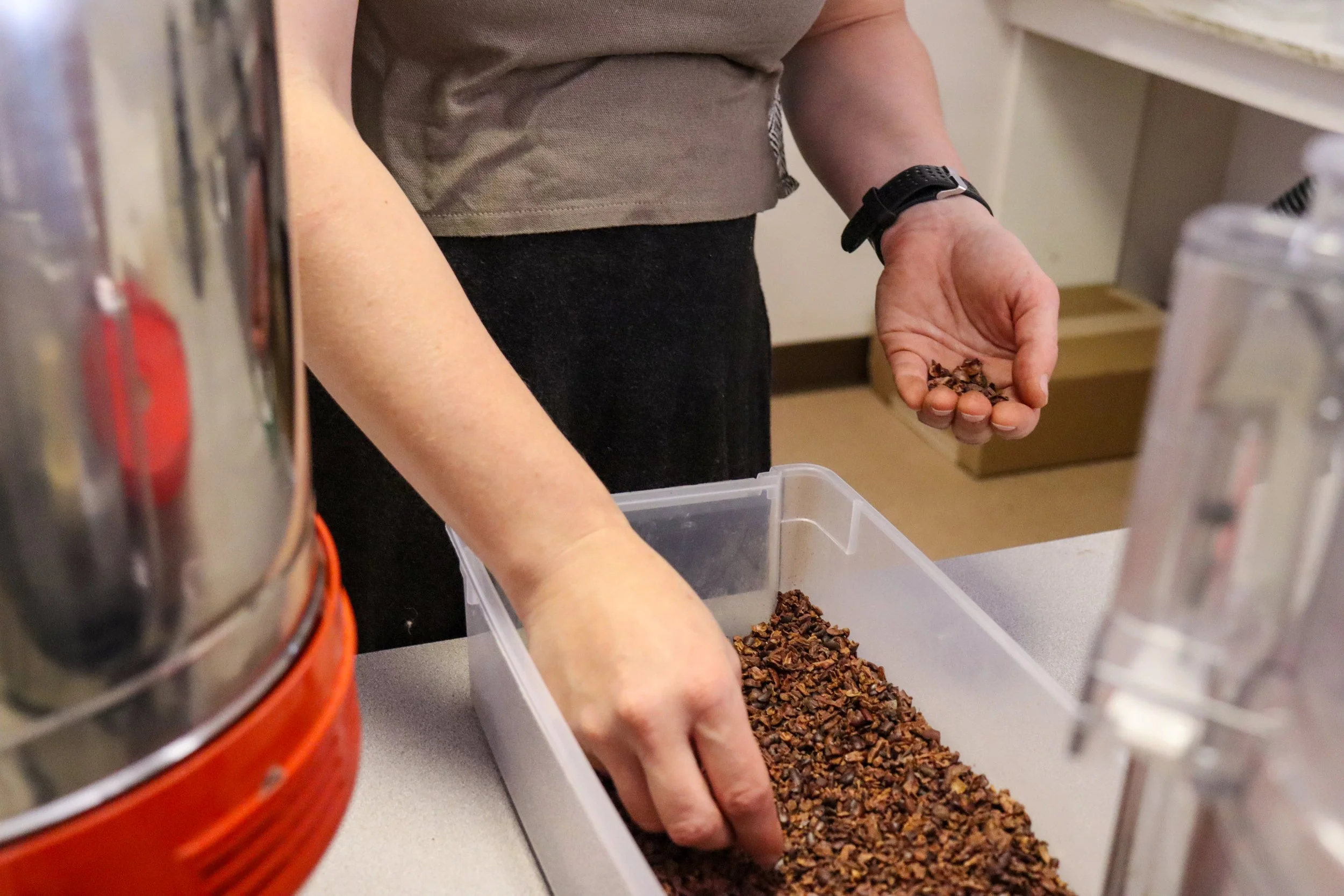 A person handling cocoa nibs in a plastic container on a countertop, with a metal container and a small appliance visible in the background.