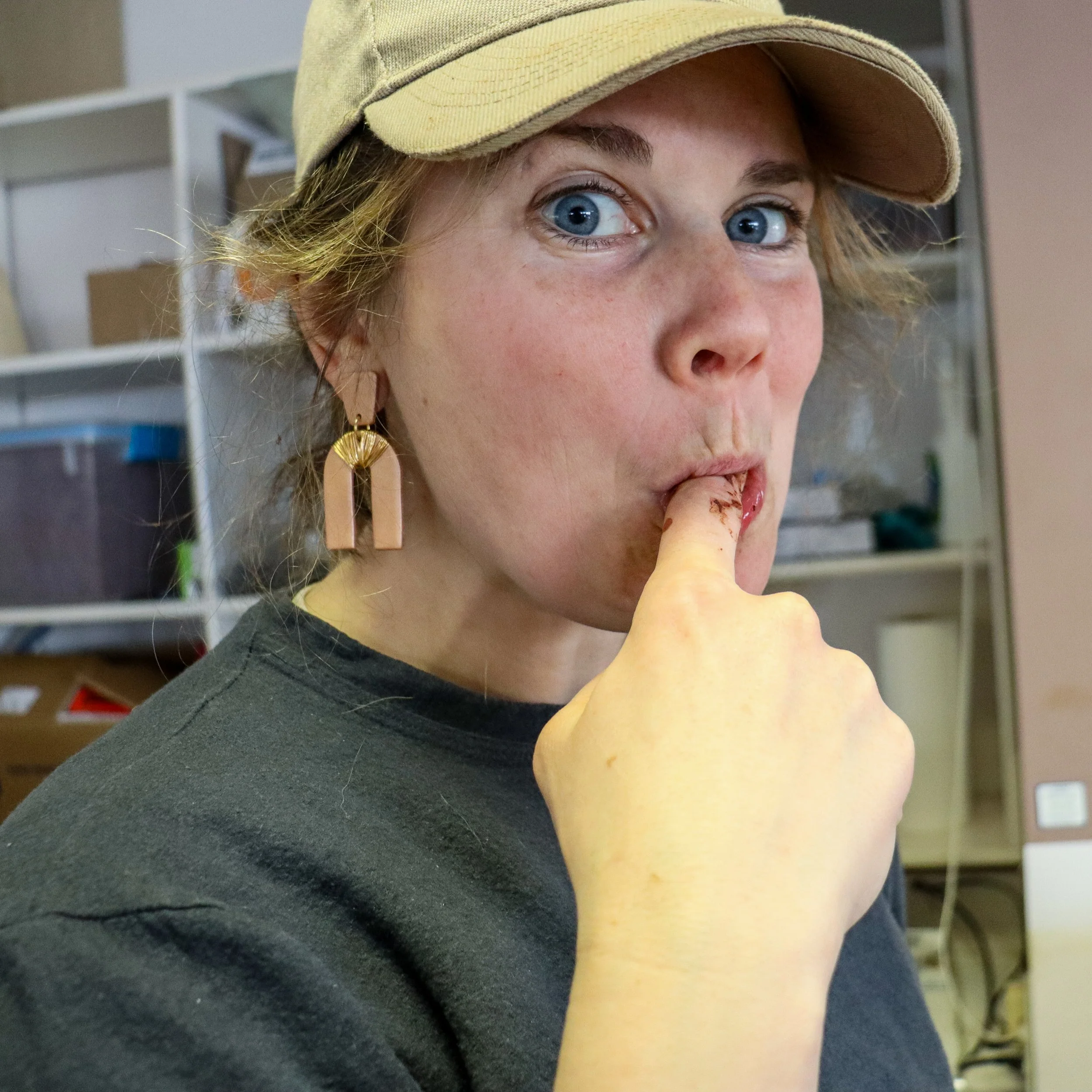 Close-up of a woman's face with blue eyes, light freckles, wearing a tan baseball cap and large pink earring, licking chocolate off of her finger.