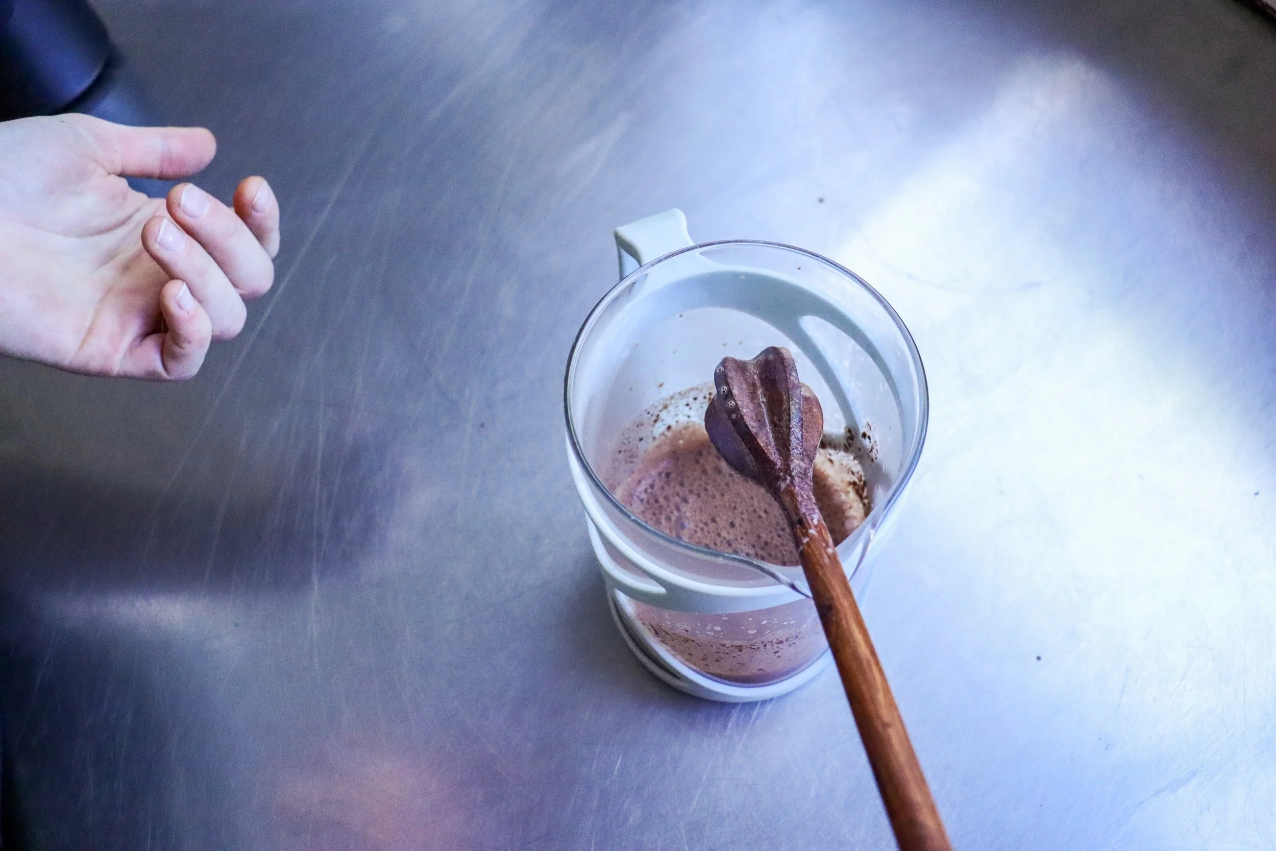 A hand holding a cocoa whisk, molinillo, over a glass mug containing a frothy, brown beverage of drinking chocolate set on a smooth metallic table.