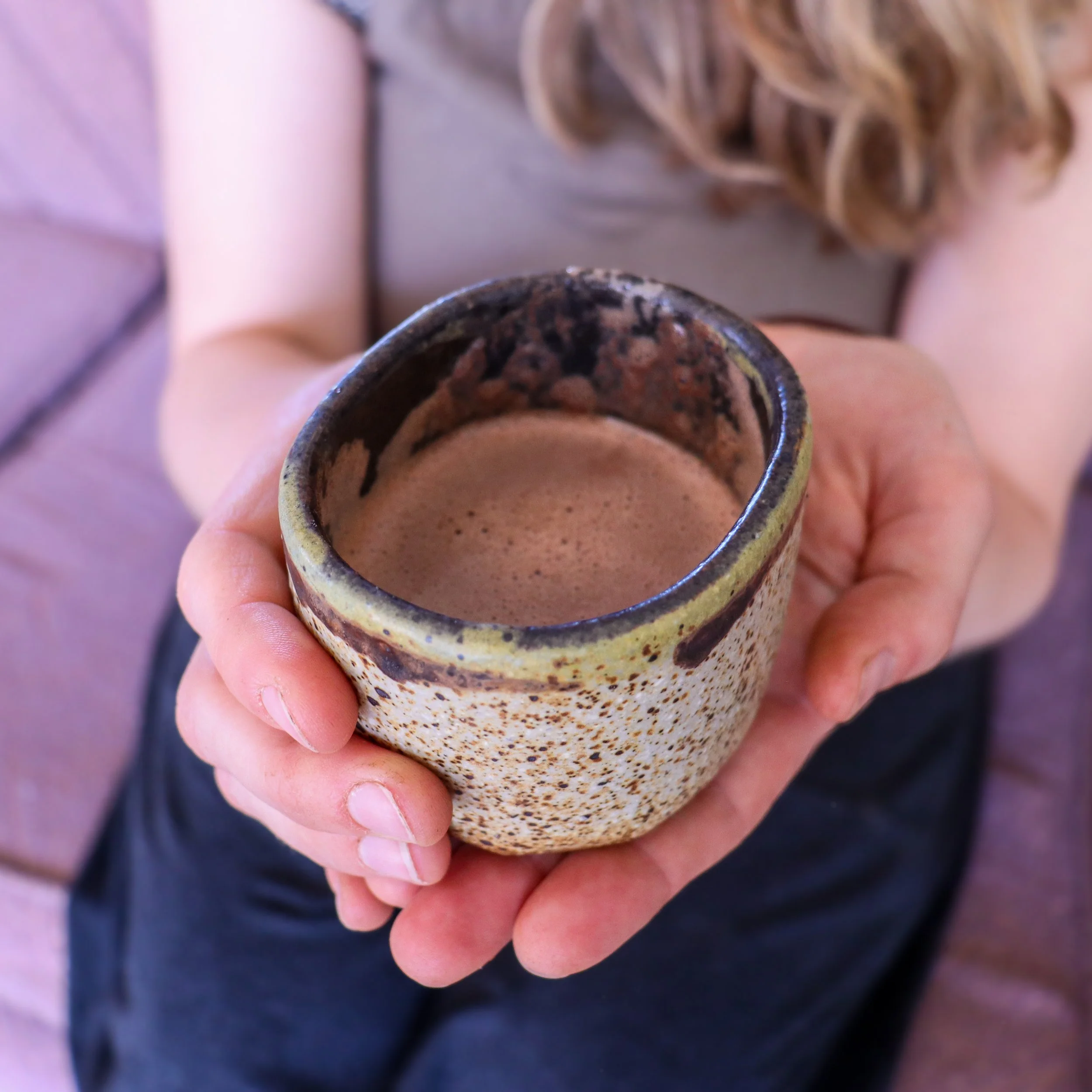 A close-up of a person's hand holding a small, glazed ceramic cup filled with artisinal drinking chocolate, with blonde wavy hair and part of a green shirt visible in the background.