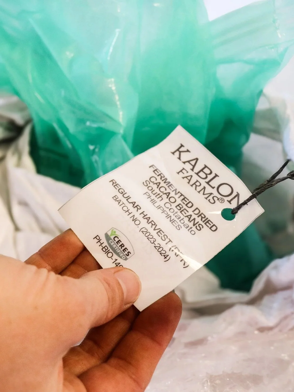 Person holding a white cocoa bean bag label with black text that reads "Kablon Farms. Feralmented Dried Cacao Beans, South Cotabato, Philippines, Regular Harvest, Batch No. 12023-2024,"