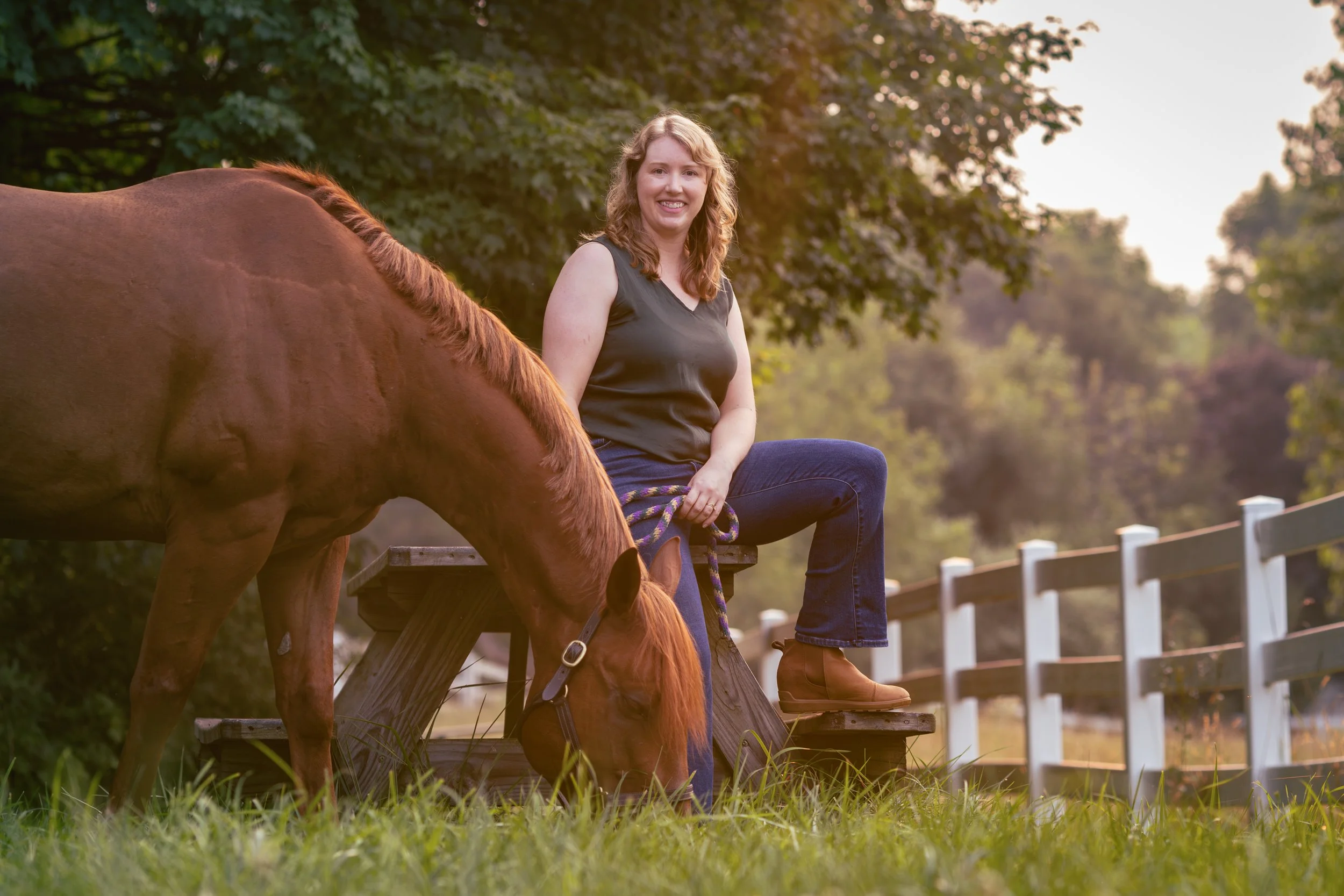 A sunset photo of a young woman sitting on a picnic table next to a chestnut horse, grazing on grass, with a background of trees and a white fence.