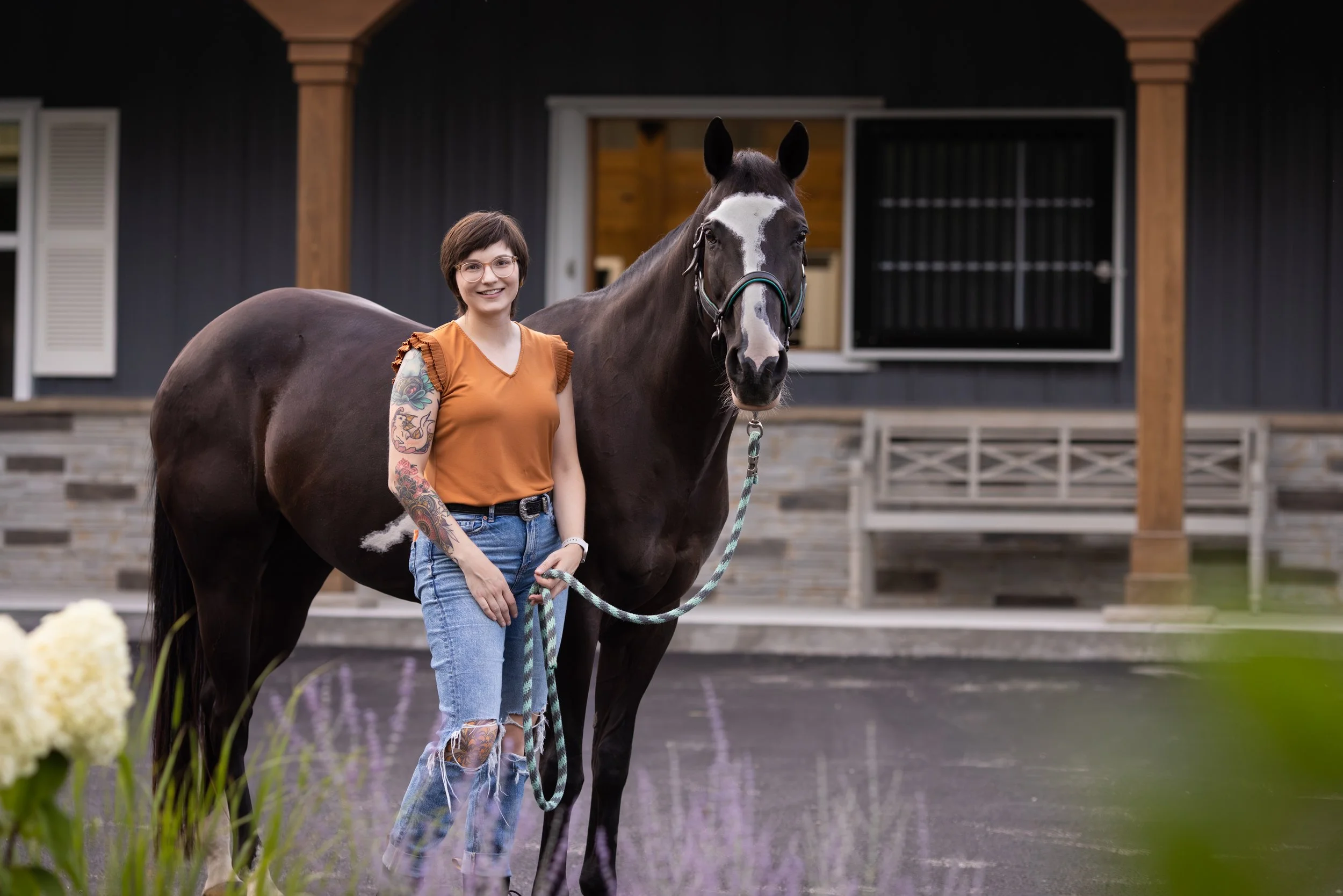 A woman with tattoos wearing an orange top and ripped jeans standing next to a black and white horse outside a barn with a dark exterior and stone accents.