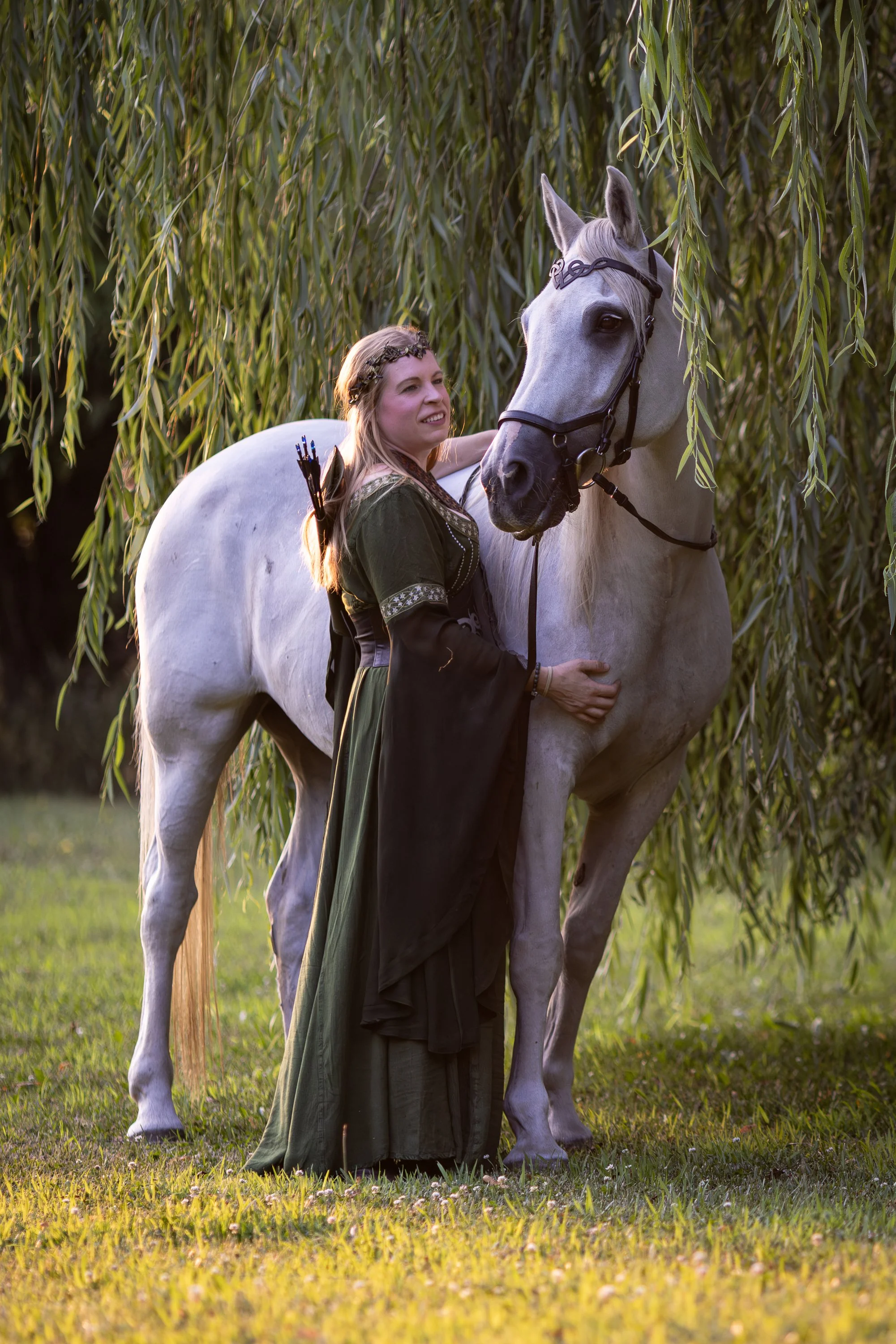 A woman dressed in a medieval-style green dress stands next to a white horse under a willow tree in a grassy field, with her hand on the horse's neck.