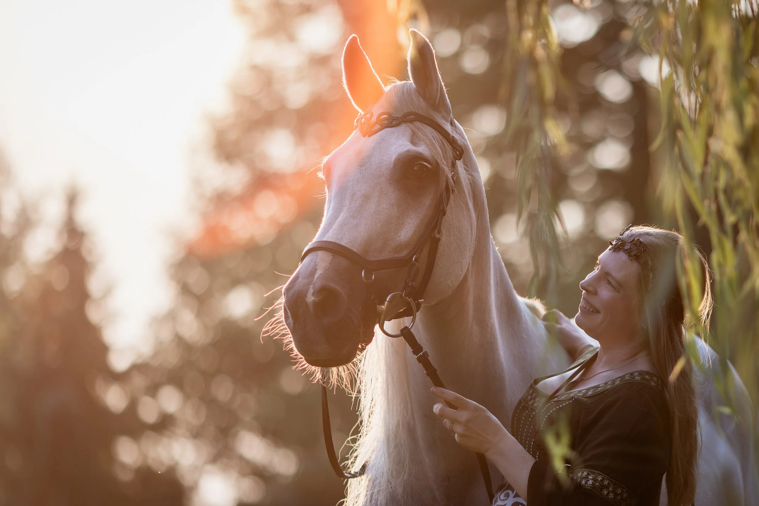 A woman smiling and petting a white horse during sunset, with trees in the background.