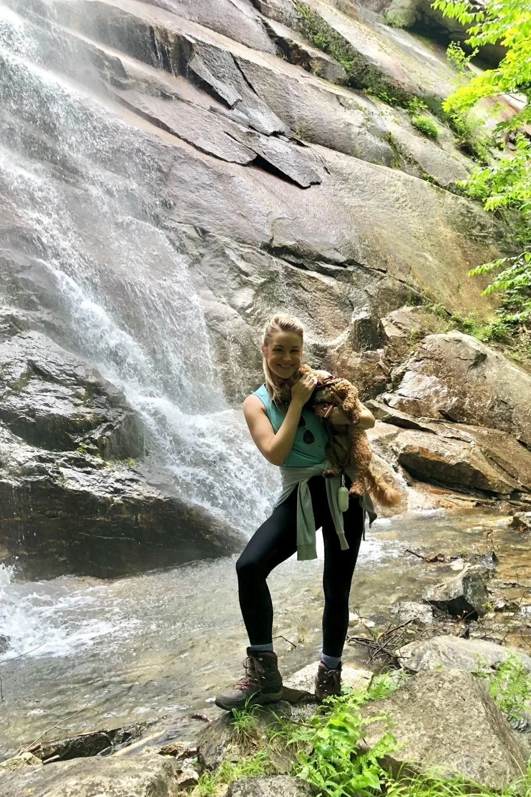 A woman is standing in front of a waterfall holding a small dog. She is smiling, wearing a teal tank top, black leggings, and hiking boots, with a gray sweatshirt tied around her waist. Green foliage surrounds the scene.