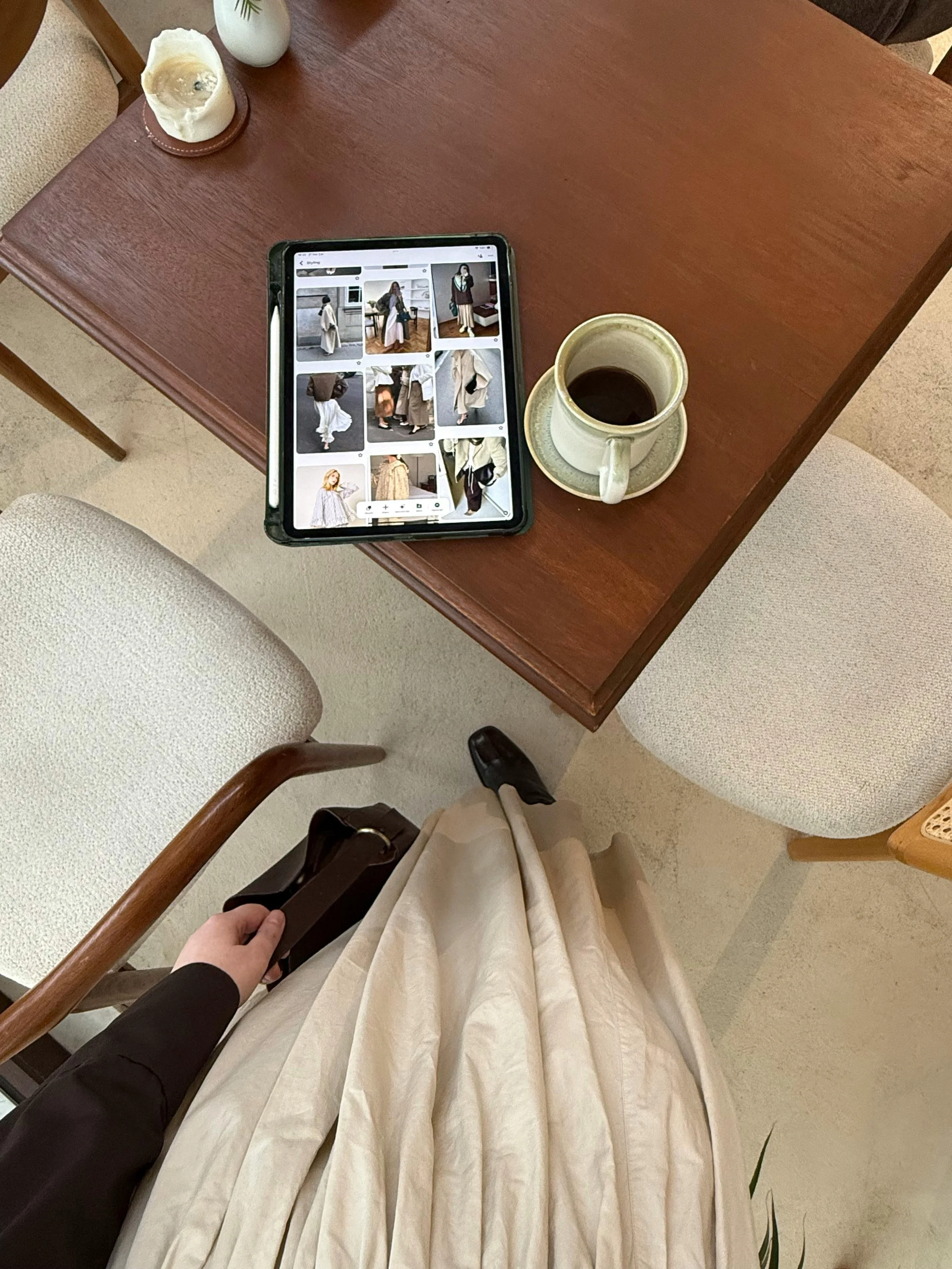 Top-down view of a person standing next to a wooden dining table, holding a purse, with a tablet showing fashion photos and a coffee cup on the table.