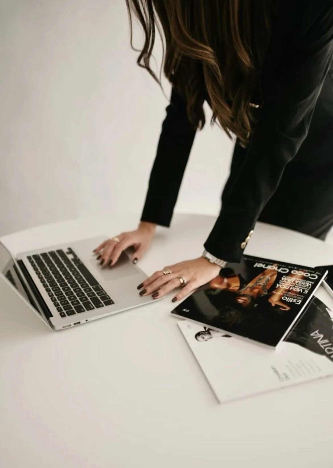 Woman with long brown hair in a black blazer leaning over a laptop on a white table with magazines.