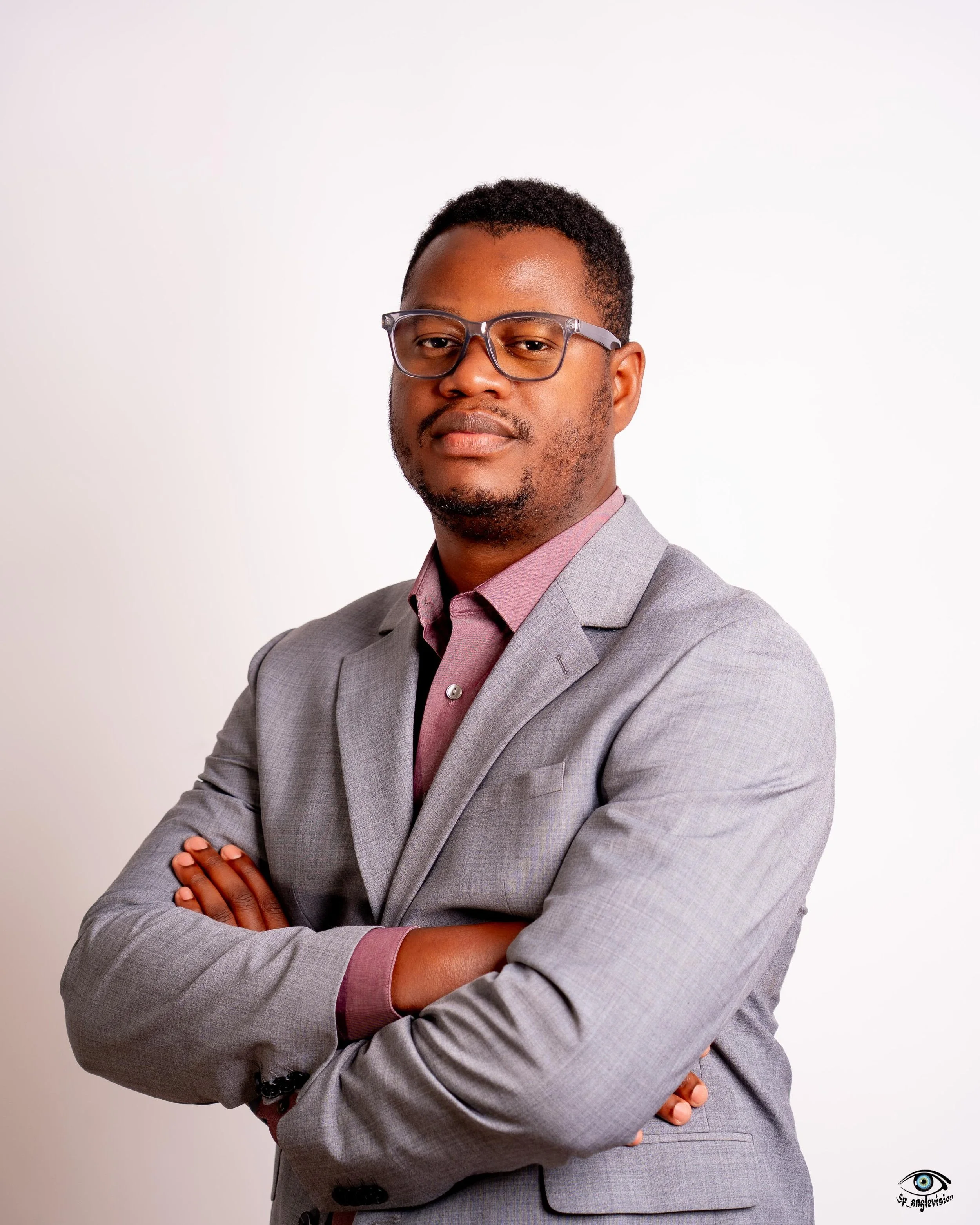 A man with glasses, wearing a light gray suit and a pink shirt, standing with arms crossed against a plain white background.