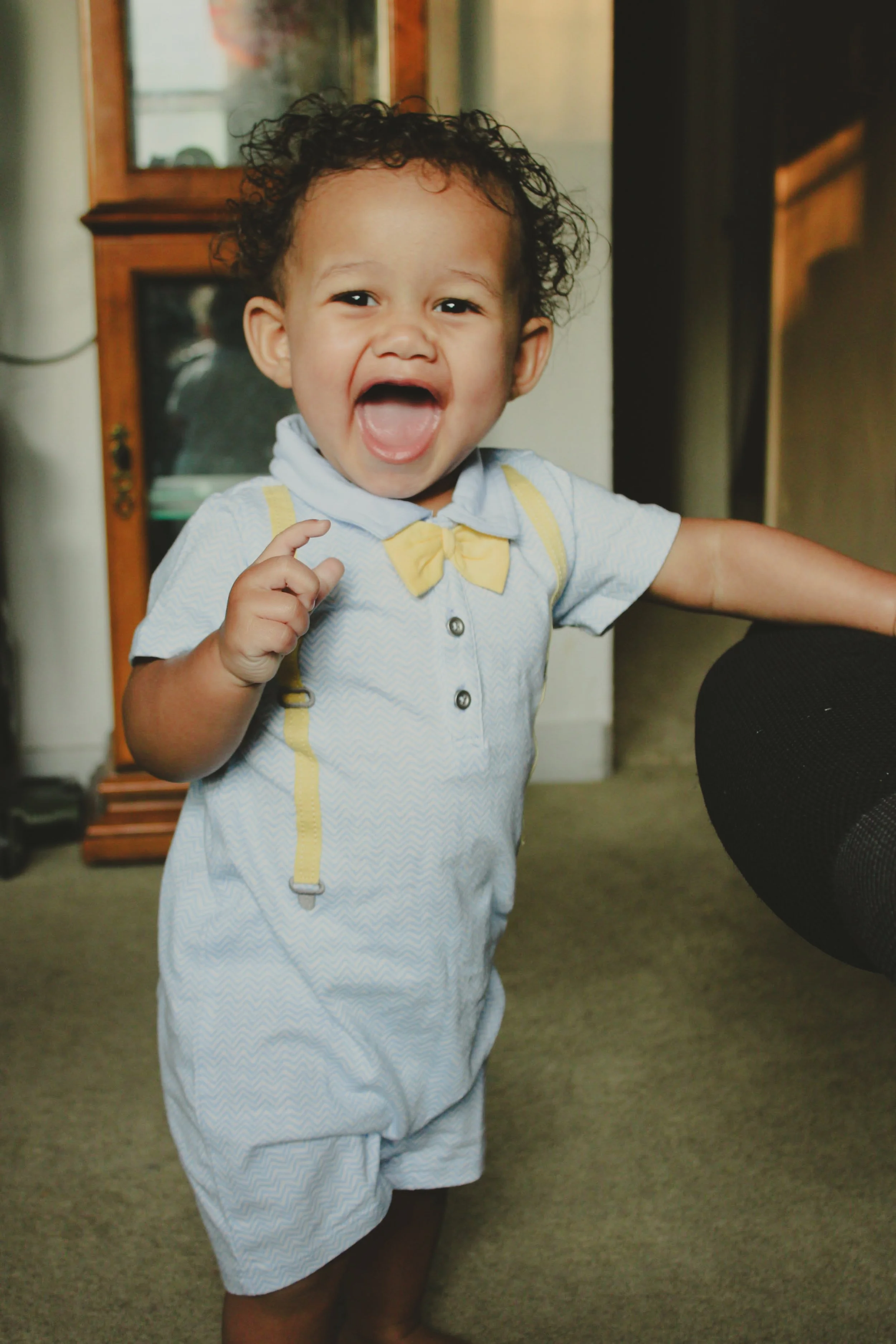 A joyful young child with curly hair smiling and pointing, dressed in a light blue outfit with a yellow bow tie, standing indoors on a carpeted floor.