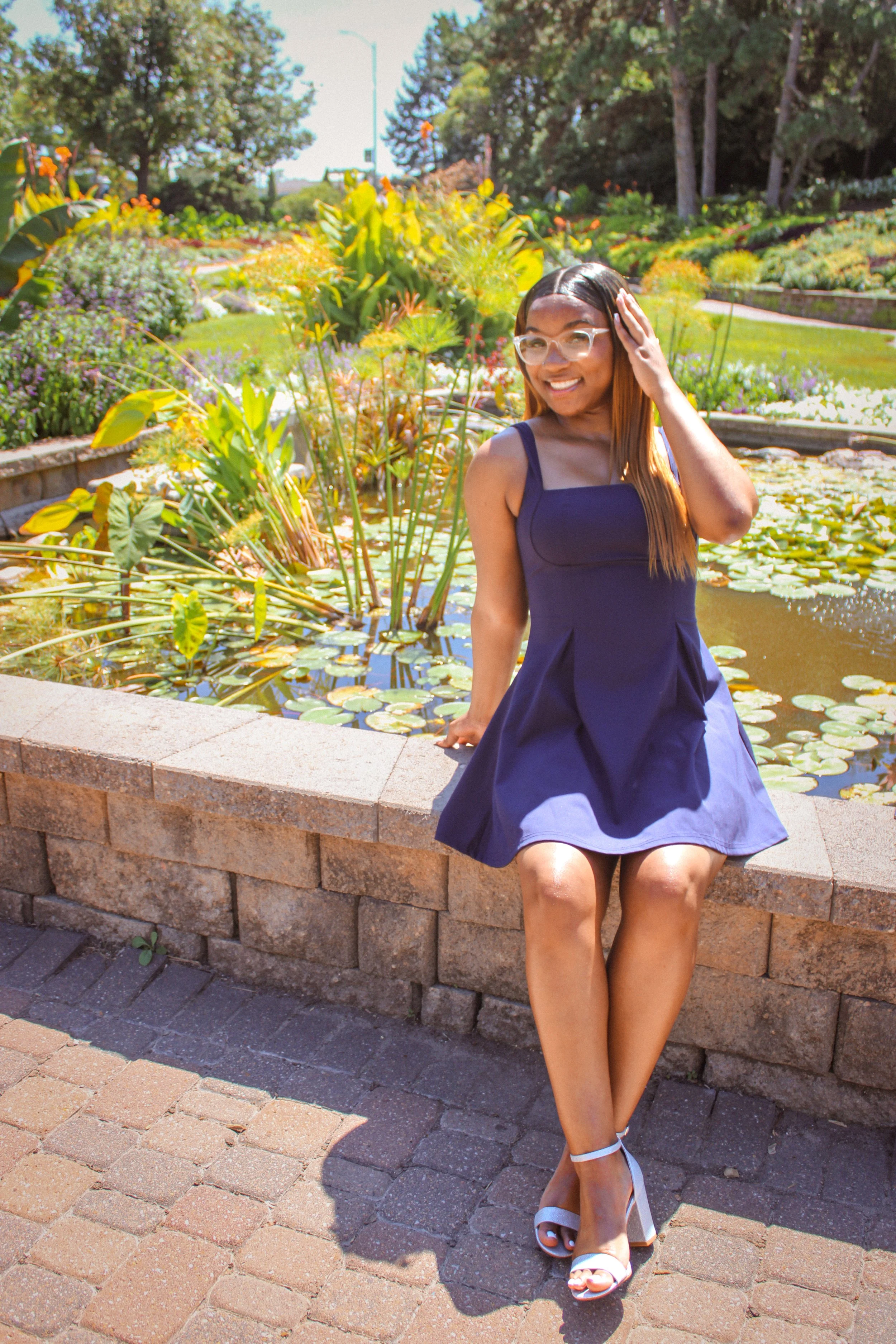 A woman wearing a navy blue dress and white high-heeled sandals sitting on a stone ledge beside a pond with lily pads, smiling and touching her hair in a garden on a sunny day.