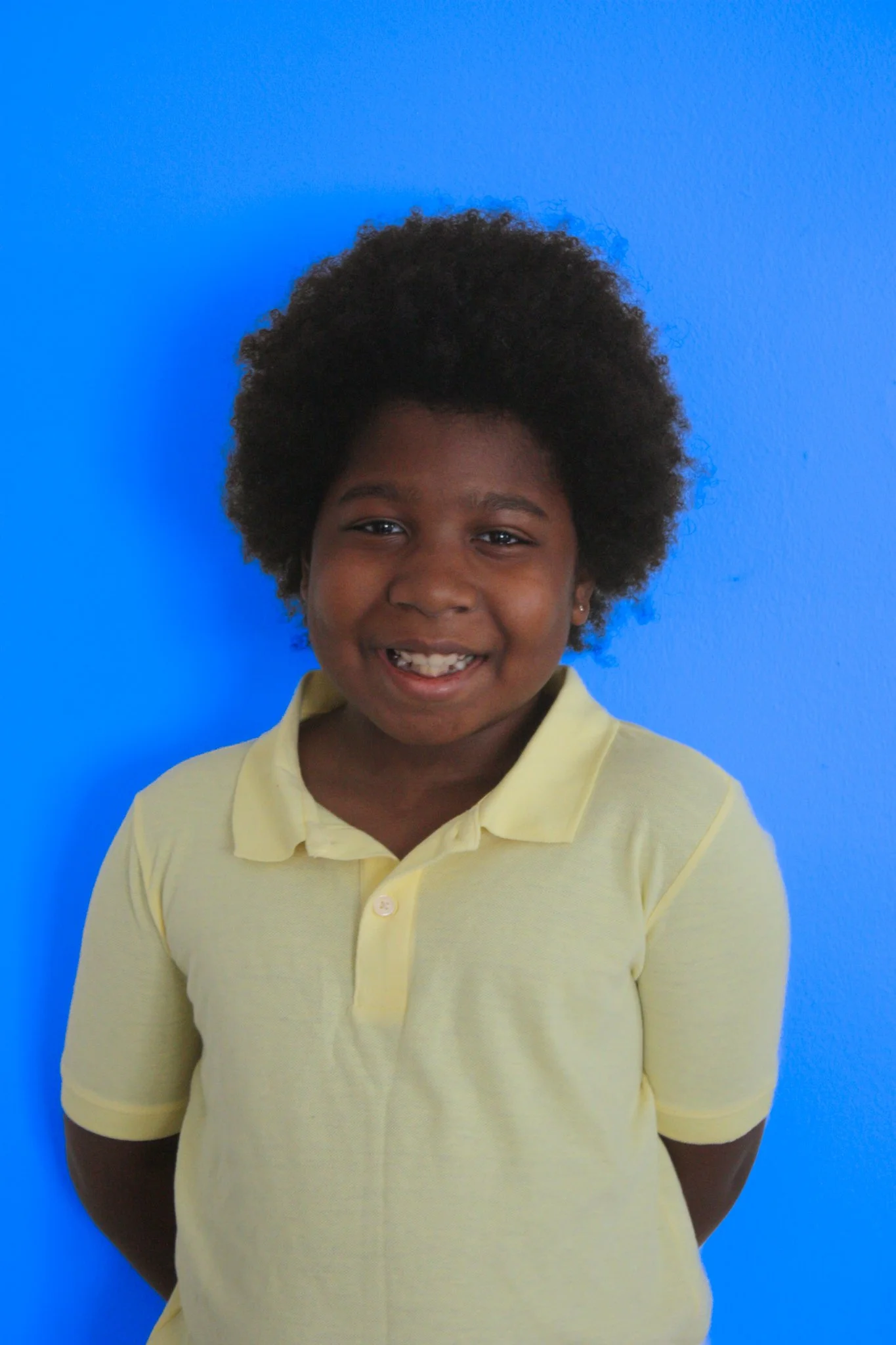Smiling young girl with curly hair wearing a yellow polo shirt stands against a bright blue wall.