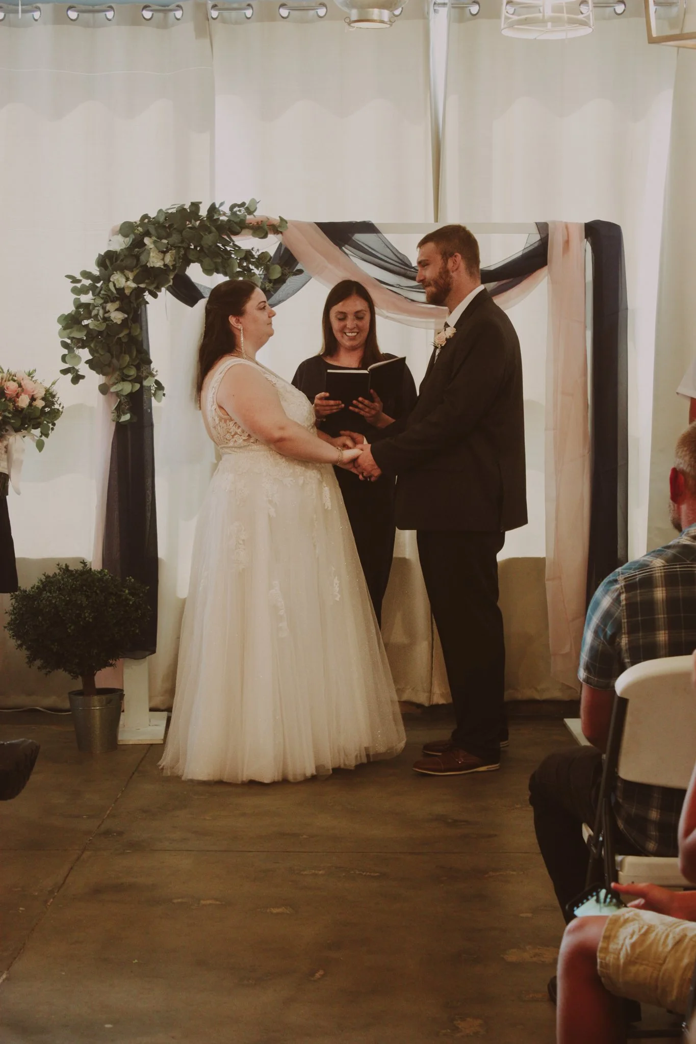 Couple holding hands during their wedding ceremony, standing in front of an officiant, with guests seated nearby, decorated with flowers and drapery.