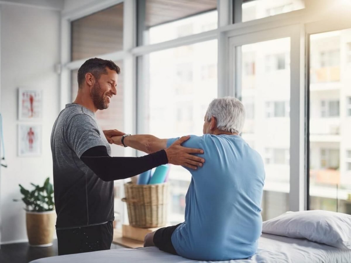 A young male physical therapist assisting an elderly male patient with shoulder exercises in a therapy room with large windows letting in natural light.