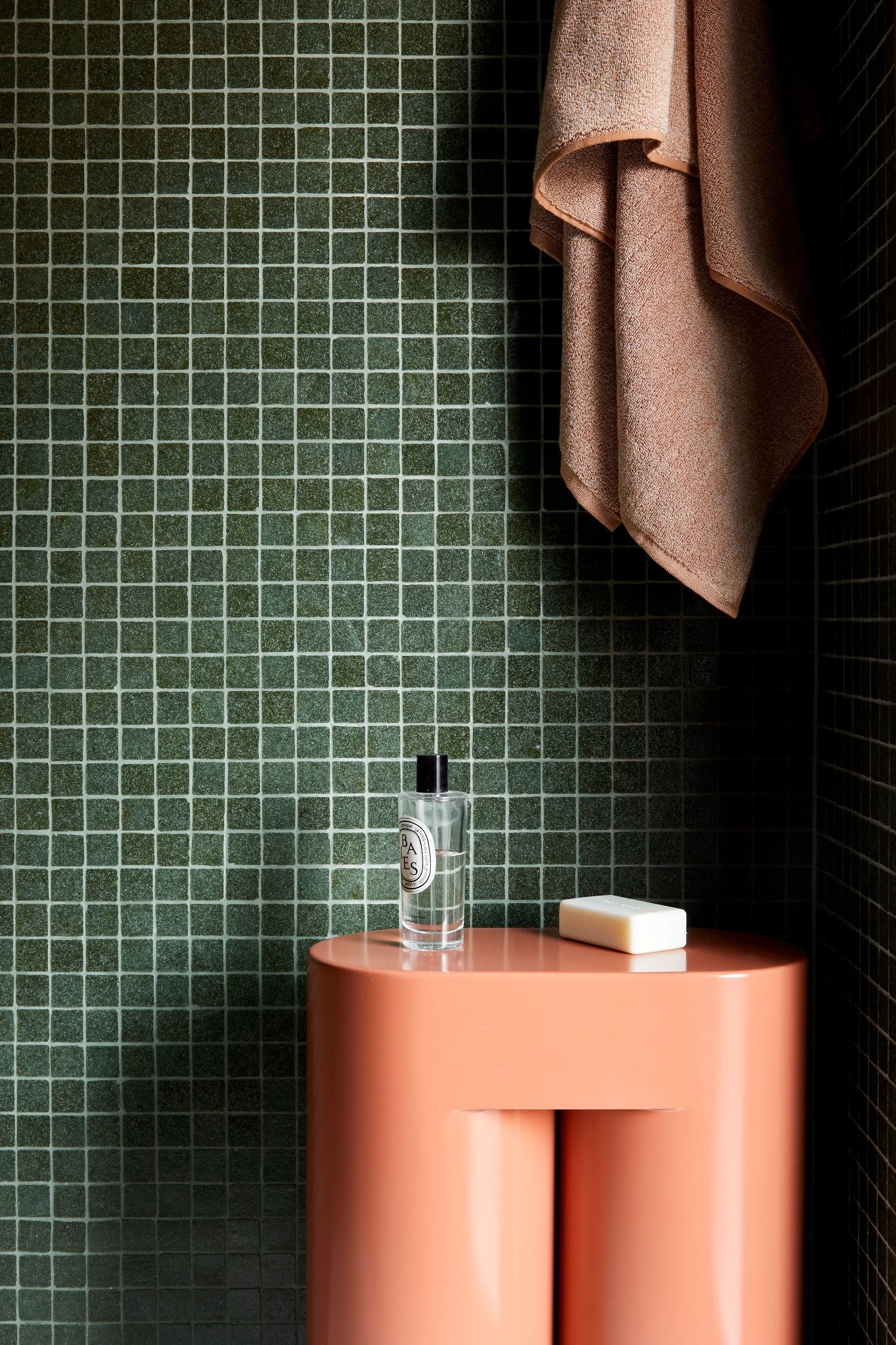 A pink round side table against a green tiled wall, with a clear bottle and soap bar on top. Beige towel hanging on the right.