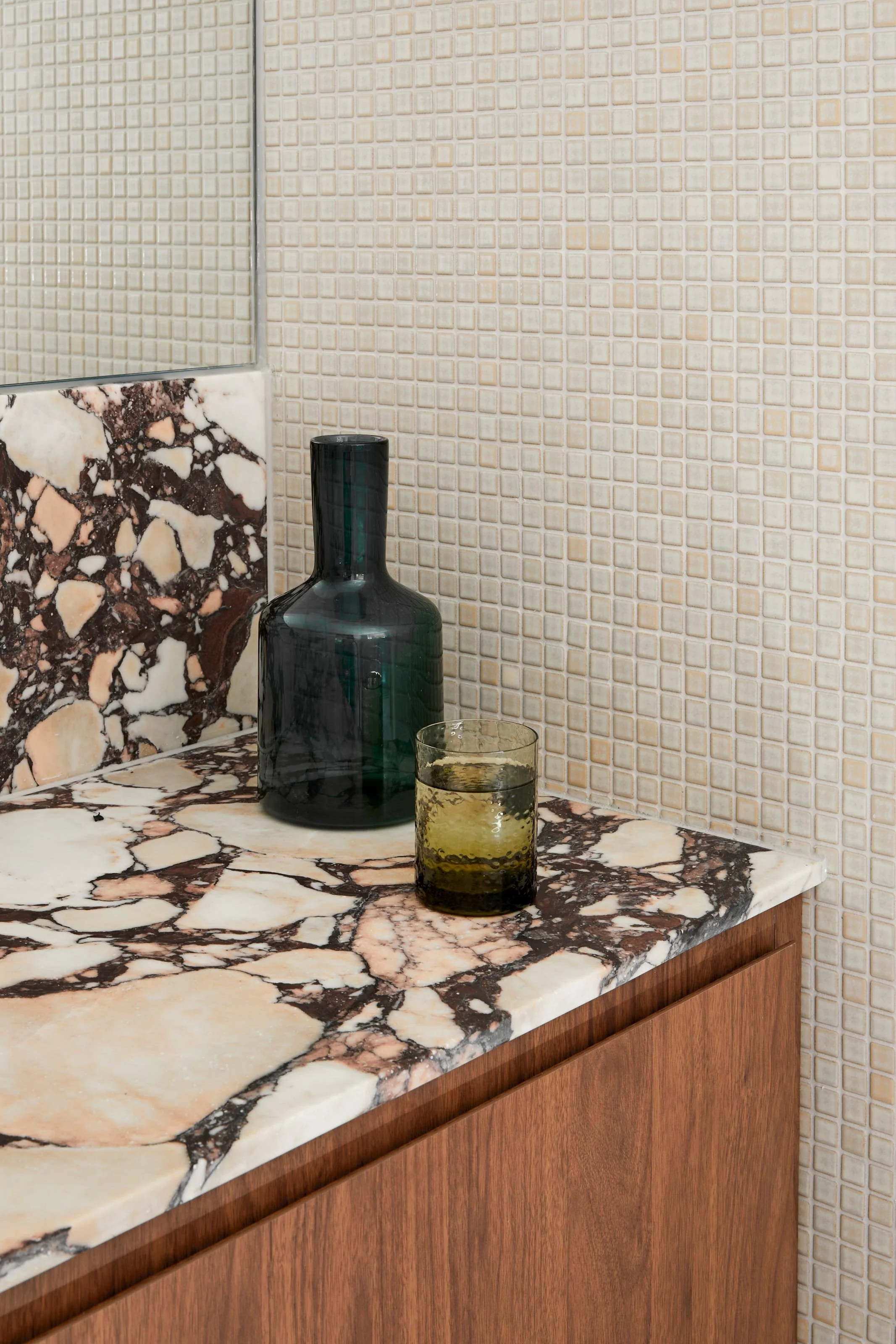 Kitchen countertop with a marble pattern, featuring a dark green glass bottle, a glass of water with lemon, and a wall with small beige mosaic tiles.