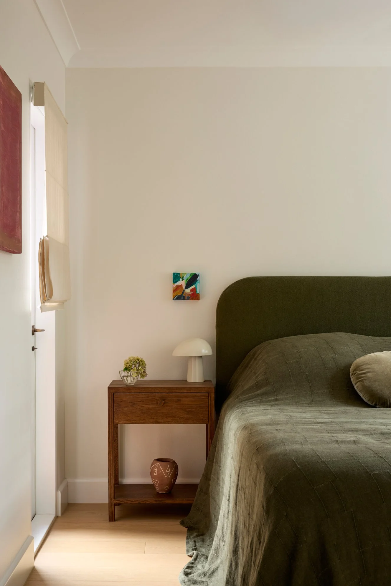 A bedroom with a bed covered in a olive green quilt, a wooden nightstand with a small lamp, a potted plant, and a decorative vase. The wall has two small colorful paintings and a larger red artwork. There is a window with a beige curtain and a fabric wall hanging.