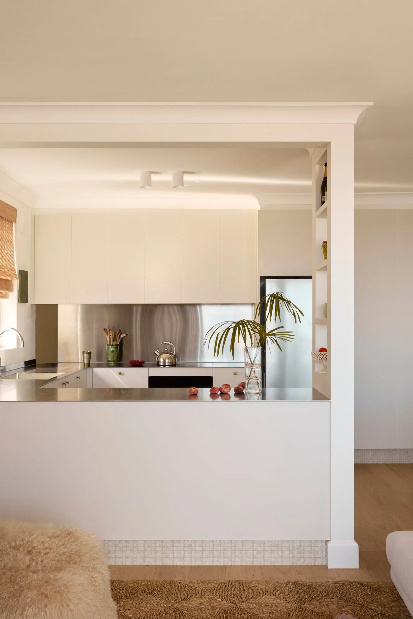 A modern white kitchen with a stainless steel countertop, white cabinets, and a potted plant on the counter. There are some peaches and a glass vase on the counter, and part of a beige textured rug and a fluffy woman's coat are visible in the foreground.