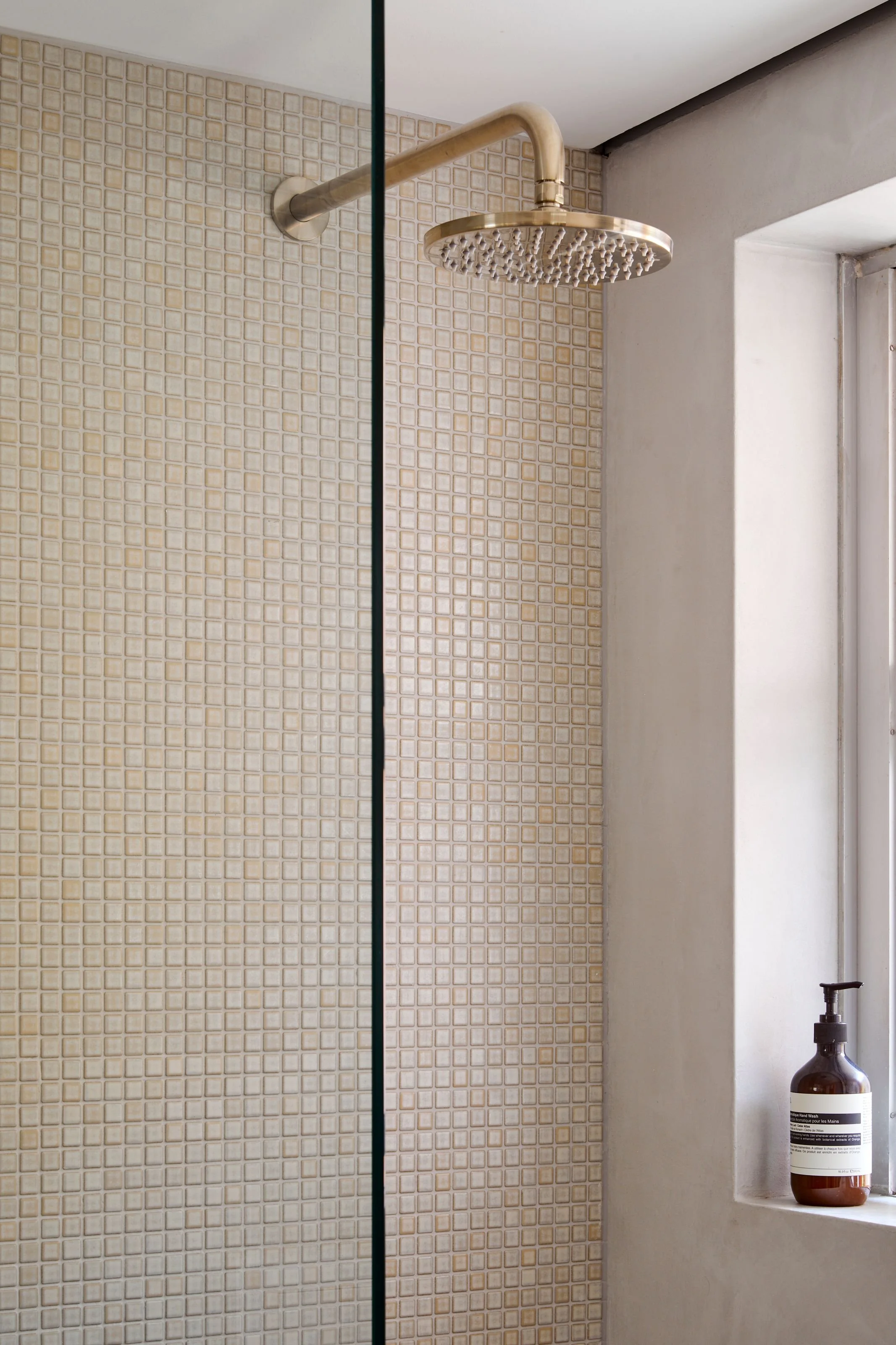 Shower area with beige mosaic tiles on the wall, a round metallic showerhead, and a place with a soap dispenser on the windowsill.
