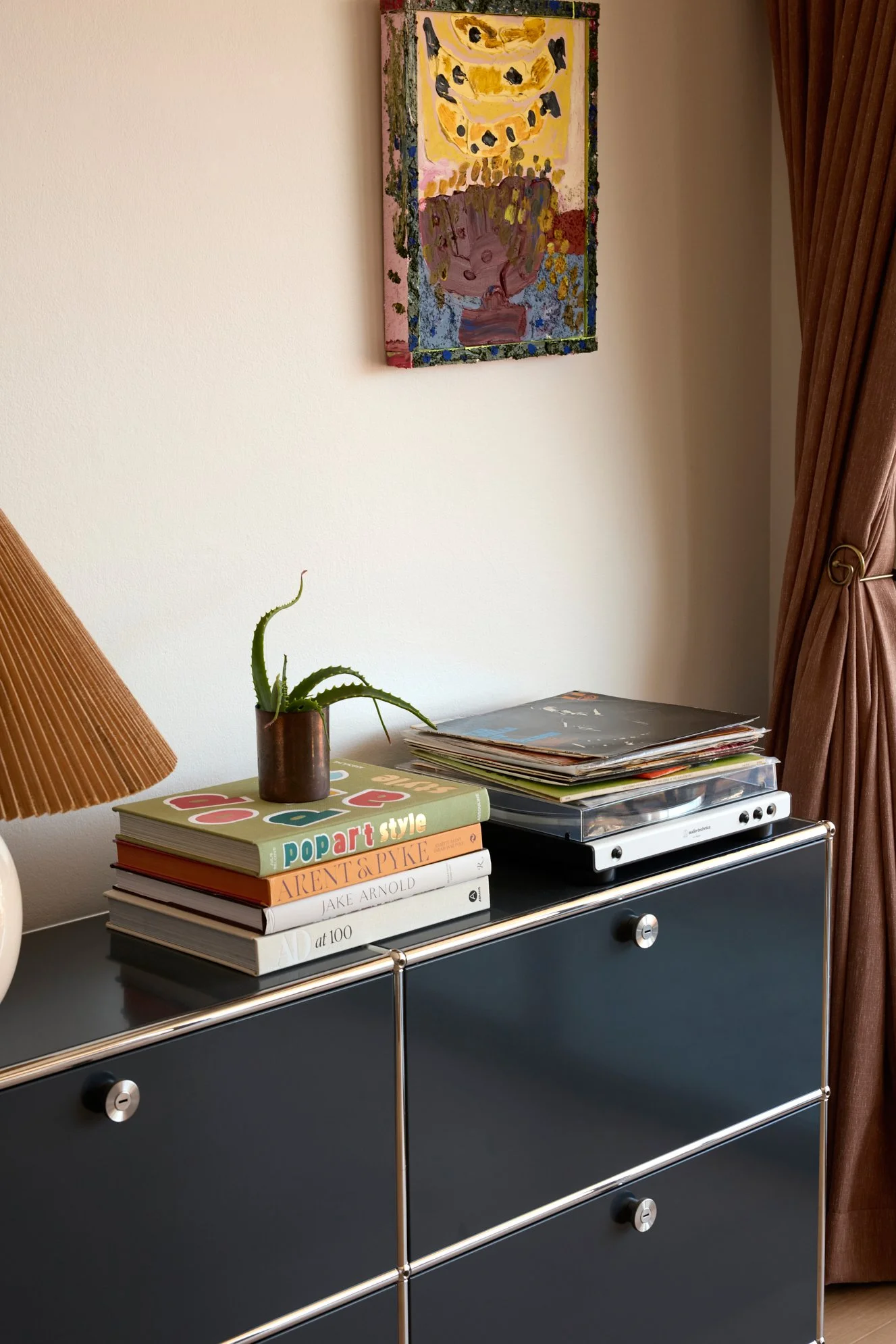 A black dresser with three drawers and silver knobs, topped with a stack of colorful art books, a small potted aloe vera plant, and a record player. A painting with abstract yellow, brown, and purple shapes hangs on the white wall behind, and a lamp with an orange shade and a brown curtain are on either side.