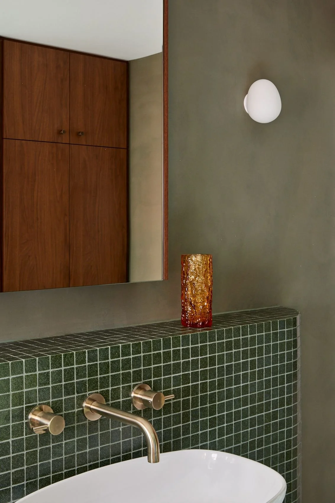 Bathroom with green tiled wall, oversize mirror, wooden cabinet, white wall-mounted light, decorative amber-colored vase, and a white bathtub with brass faucet.