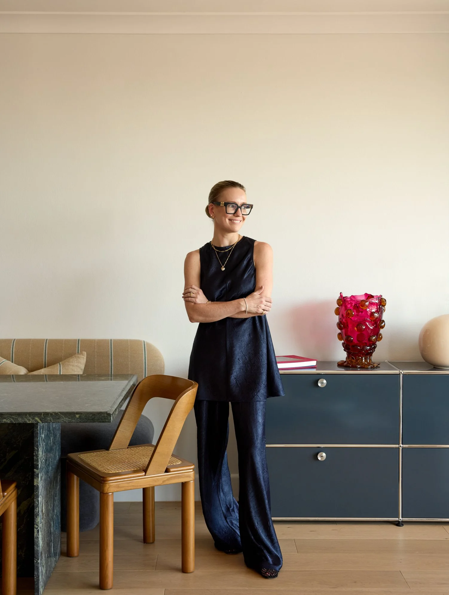 A woman in a black sleeveless dress, large black glasses, and layered necklaces stands with arms crossed, smiling in a modern interior with a beige wall, wooden chair, marble table, a blue chest of drawers, a pink decorative vase, and a beige round object.