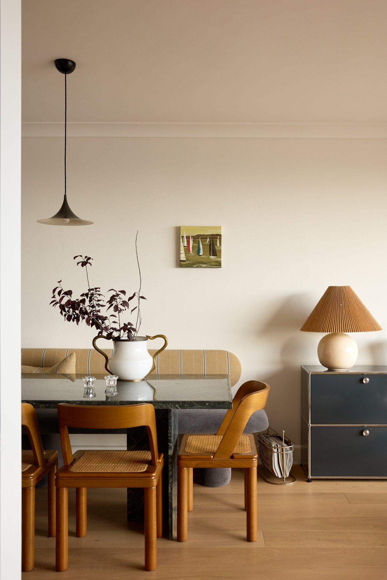 A cozy dining room with four wooden chairs around a black marble table, a white vase with reddish-purple branches, a vintage beige and blue cabinet, a table lamp with a brown lampshade, a small abstract painting on the wall, and a glass with a small crystal object on the table.