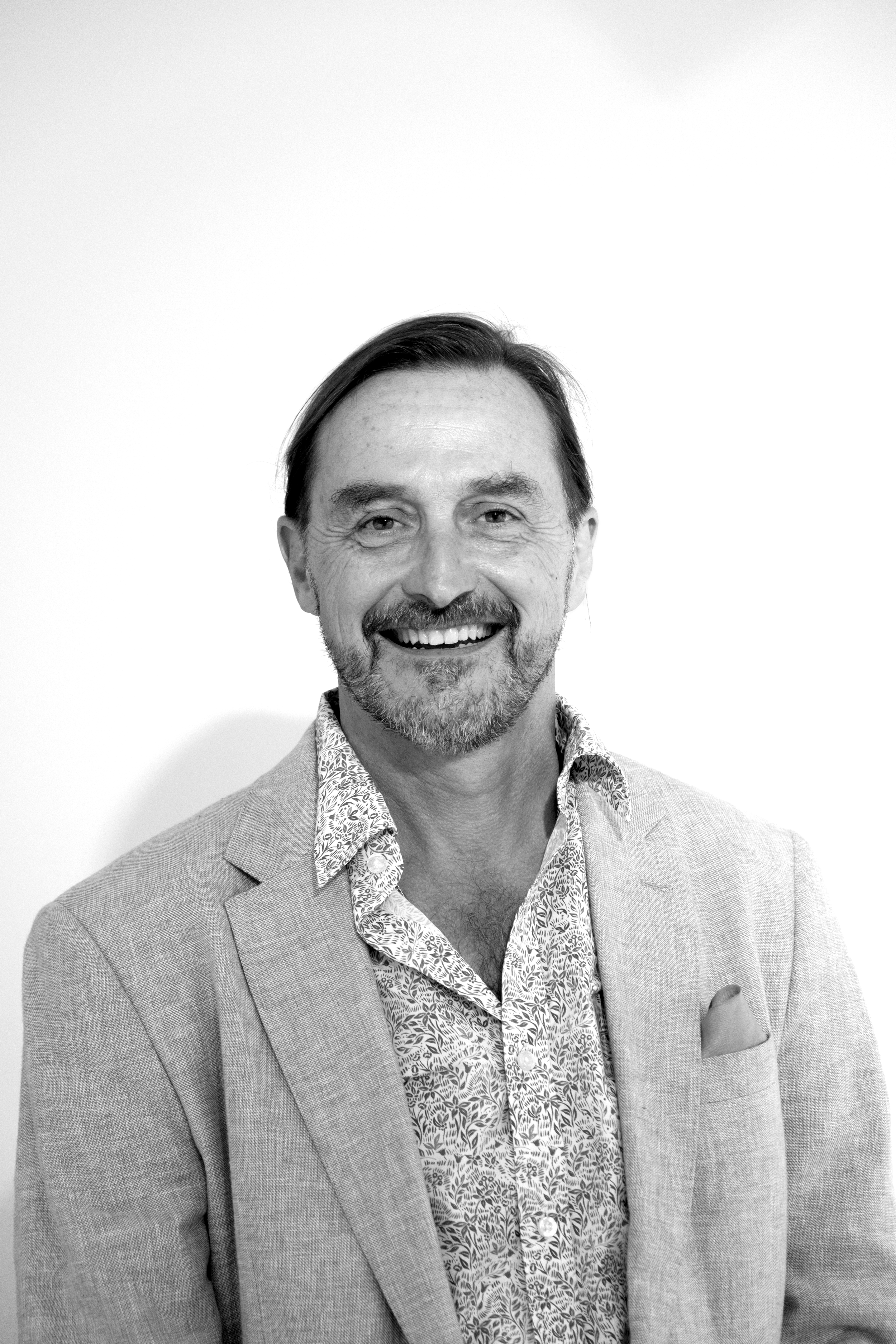 A smiling middle-aged man with a beard and short hair, wearing a patterned shirt and a light-colored blazer, standing against a plain white background.