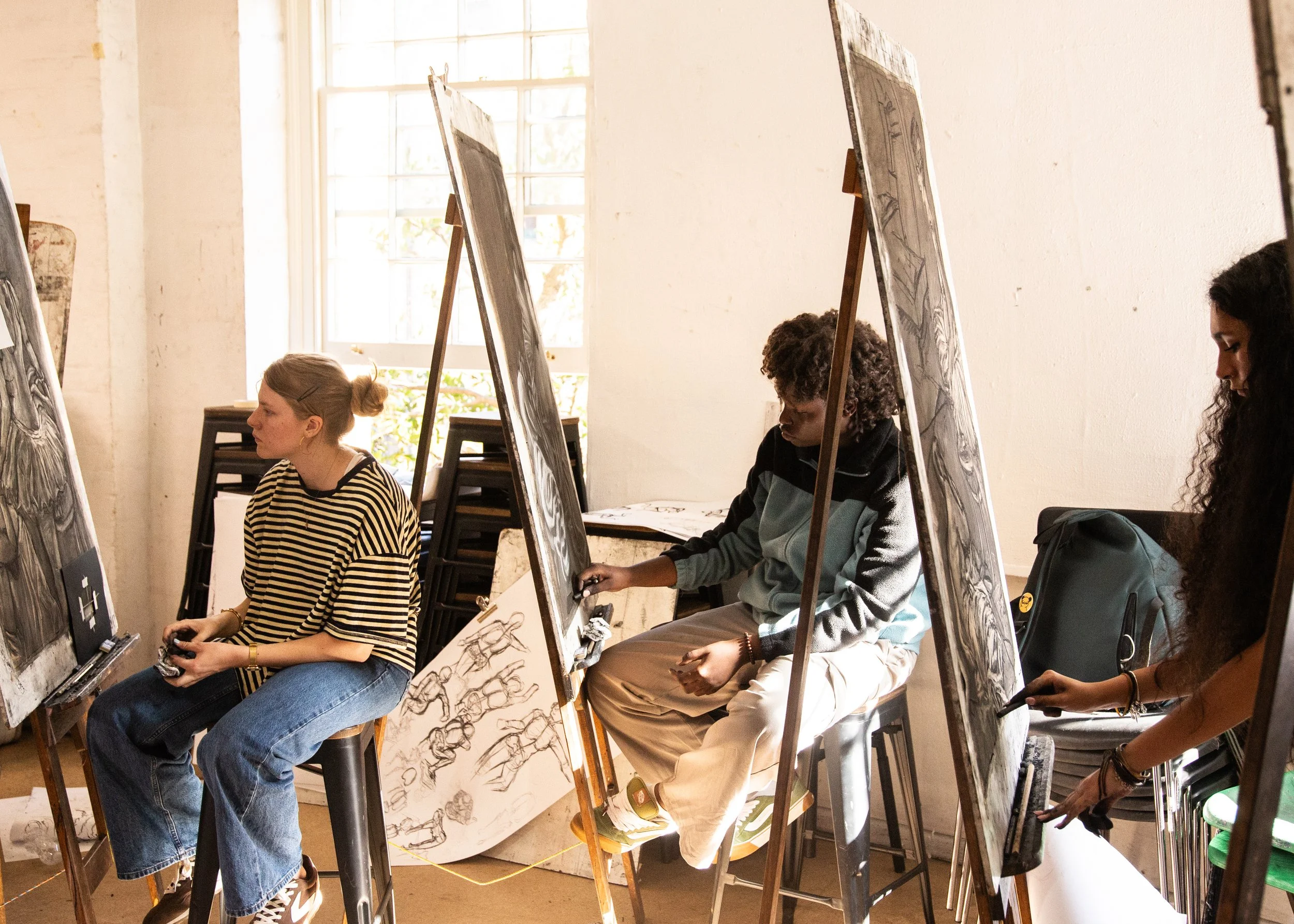 Group of people participating in a daytime drawing or art class in a bright studio with large windows, sketching on paper seated at easels.