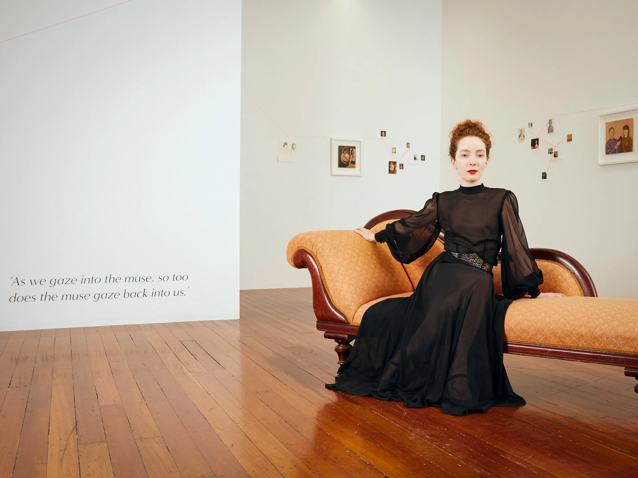 A woman with curly red hair and red lipstick is sitting on an orange vintage sofa in an art gallery. There are framed photographs and small pictures arranged in an artistic display on the white wall behind her. The floor is made of wooden planks.