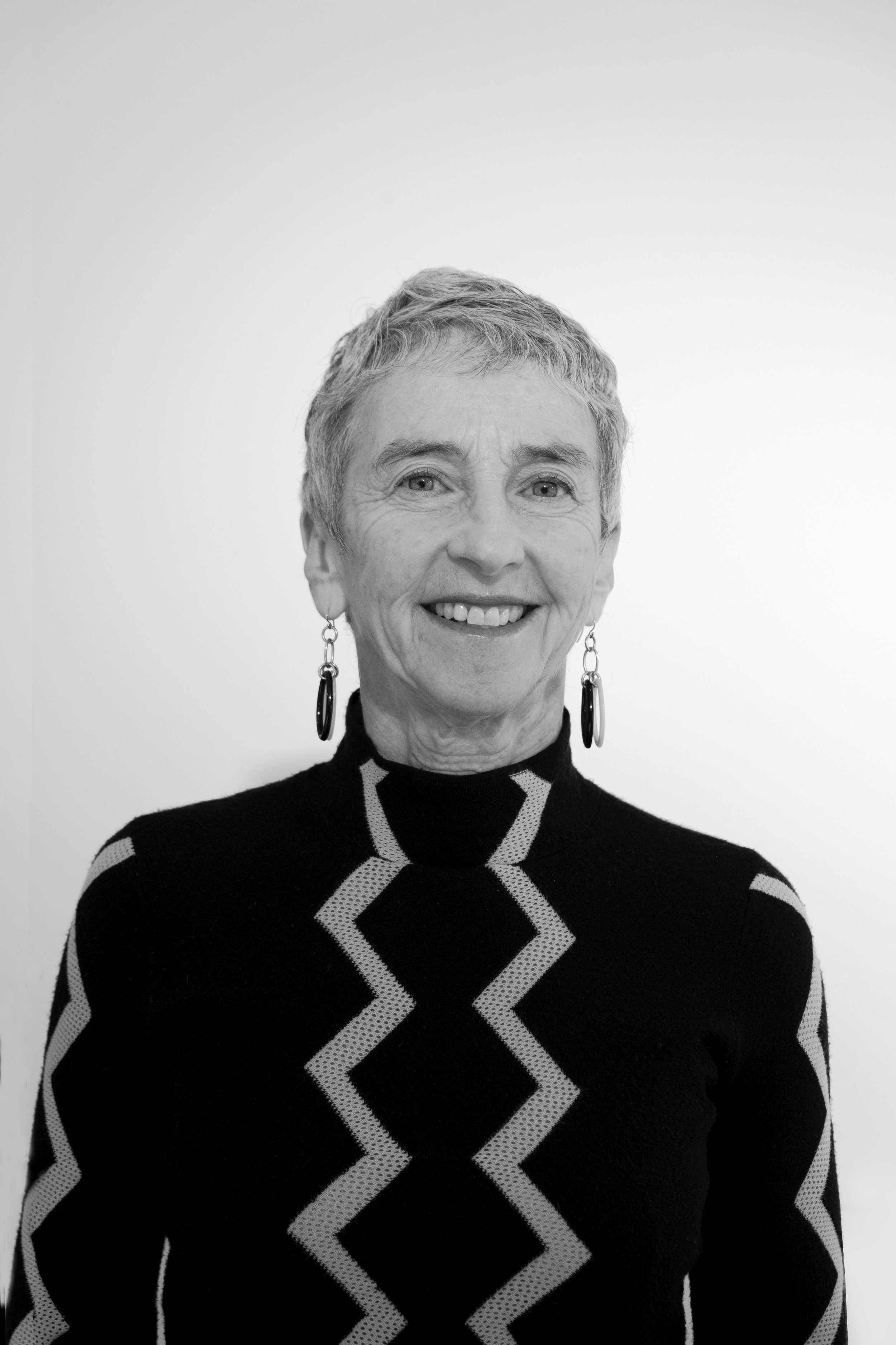 Black and white portrait of a smiling woman with short hair, wearing earrings and a patterned sweater, against a plain background.