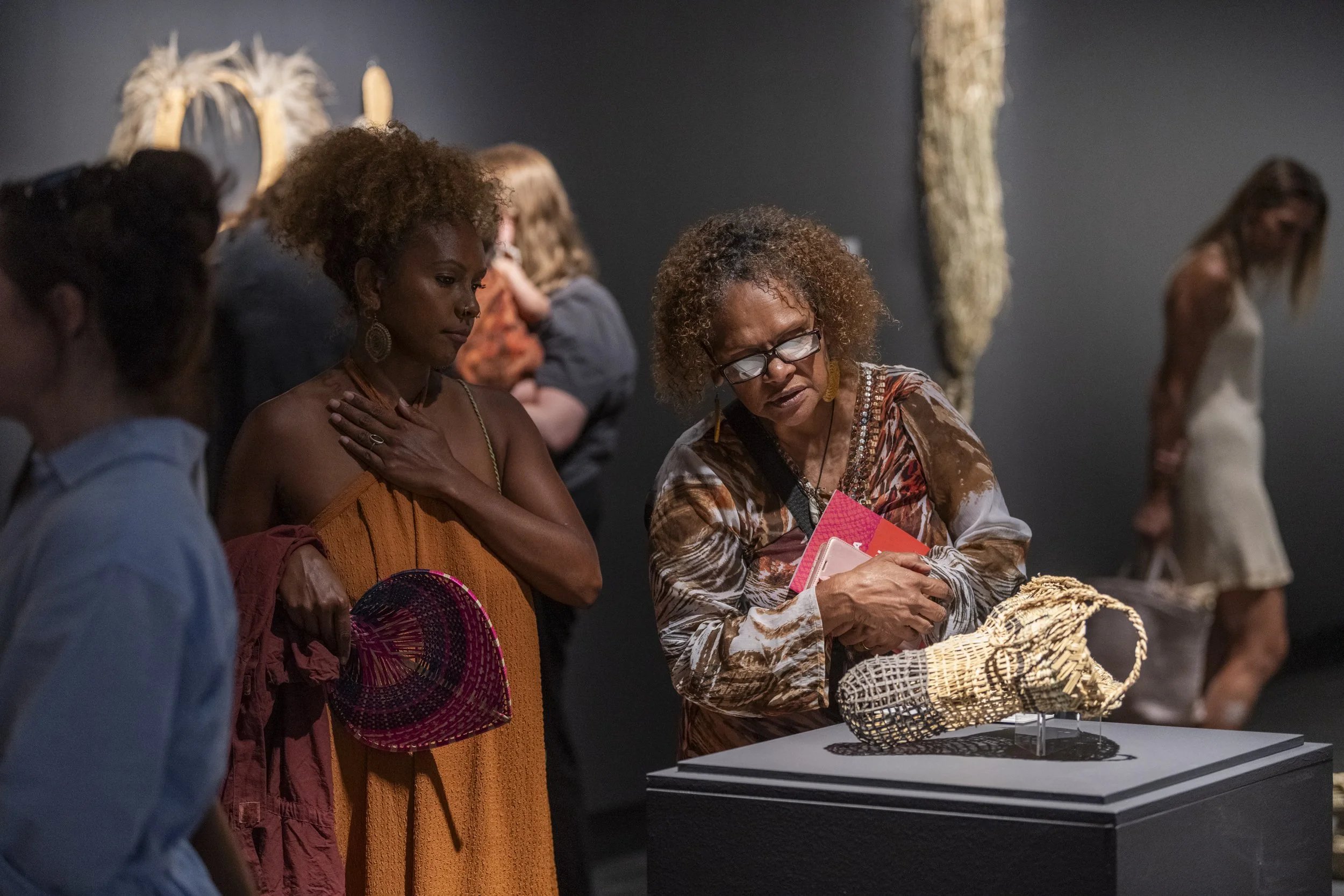A group of women in an art gallery examining a woven sculpture on display, with some standing close and focused, and others in the background.