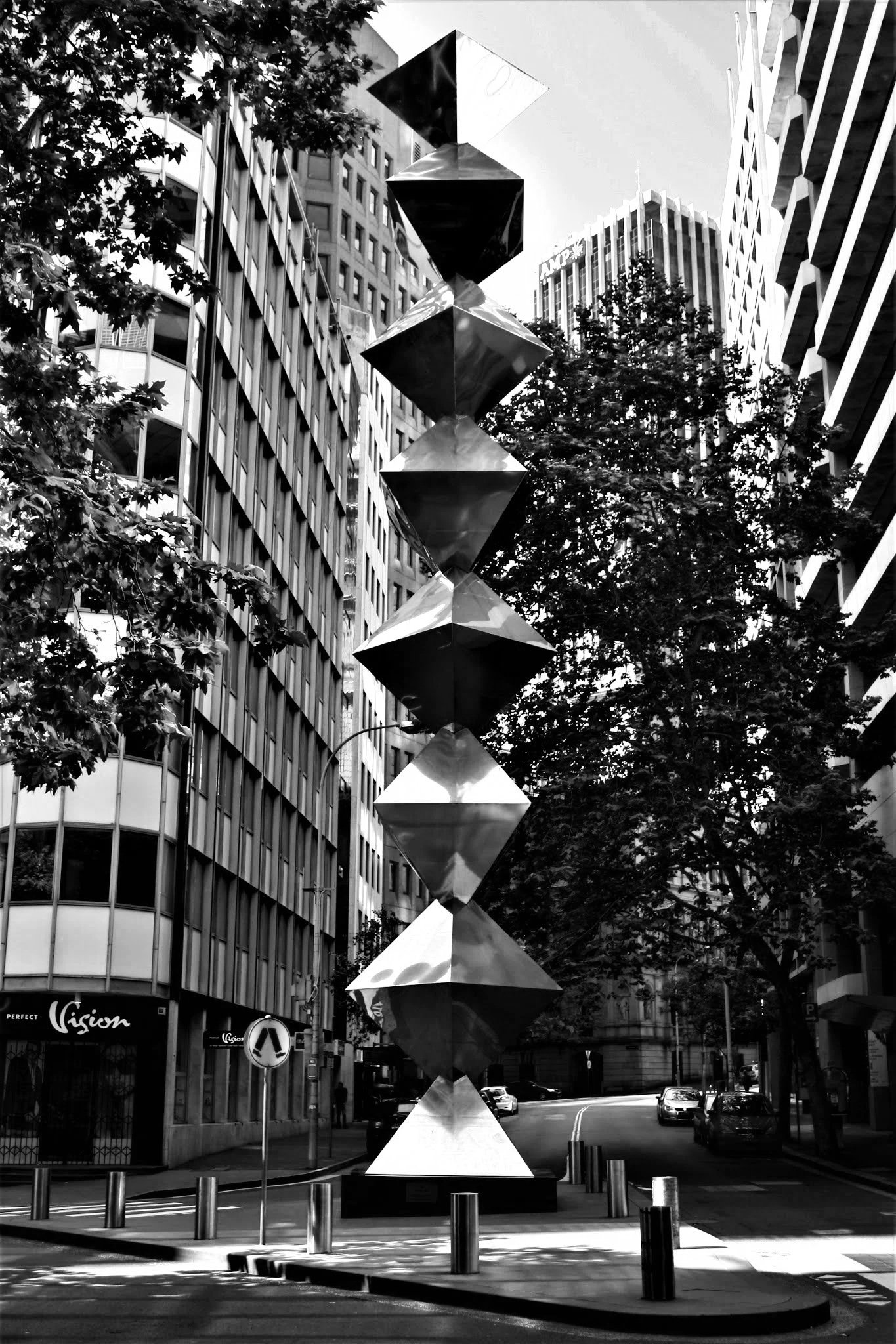 A street scene in black and white featuring modern office buildings, trees, cars parked on the street, and a tall abstract sculpture with geometric shapes in the foreground.