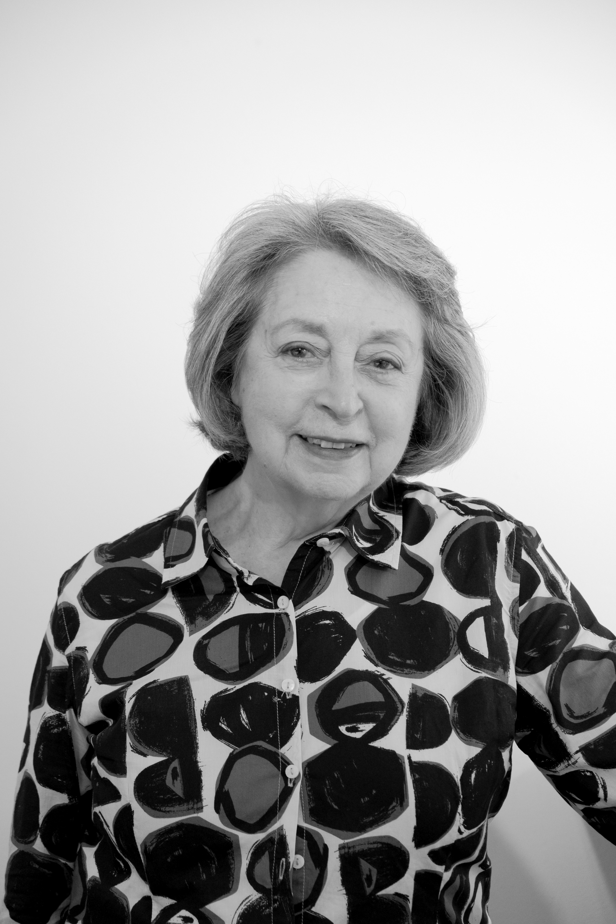 A woman with short, wavy hair smiling, wearing a patterned blouse with large abstract shapes, in front of a plain light-colored background.