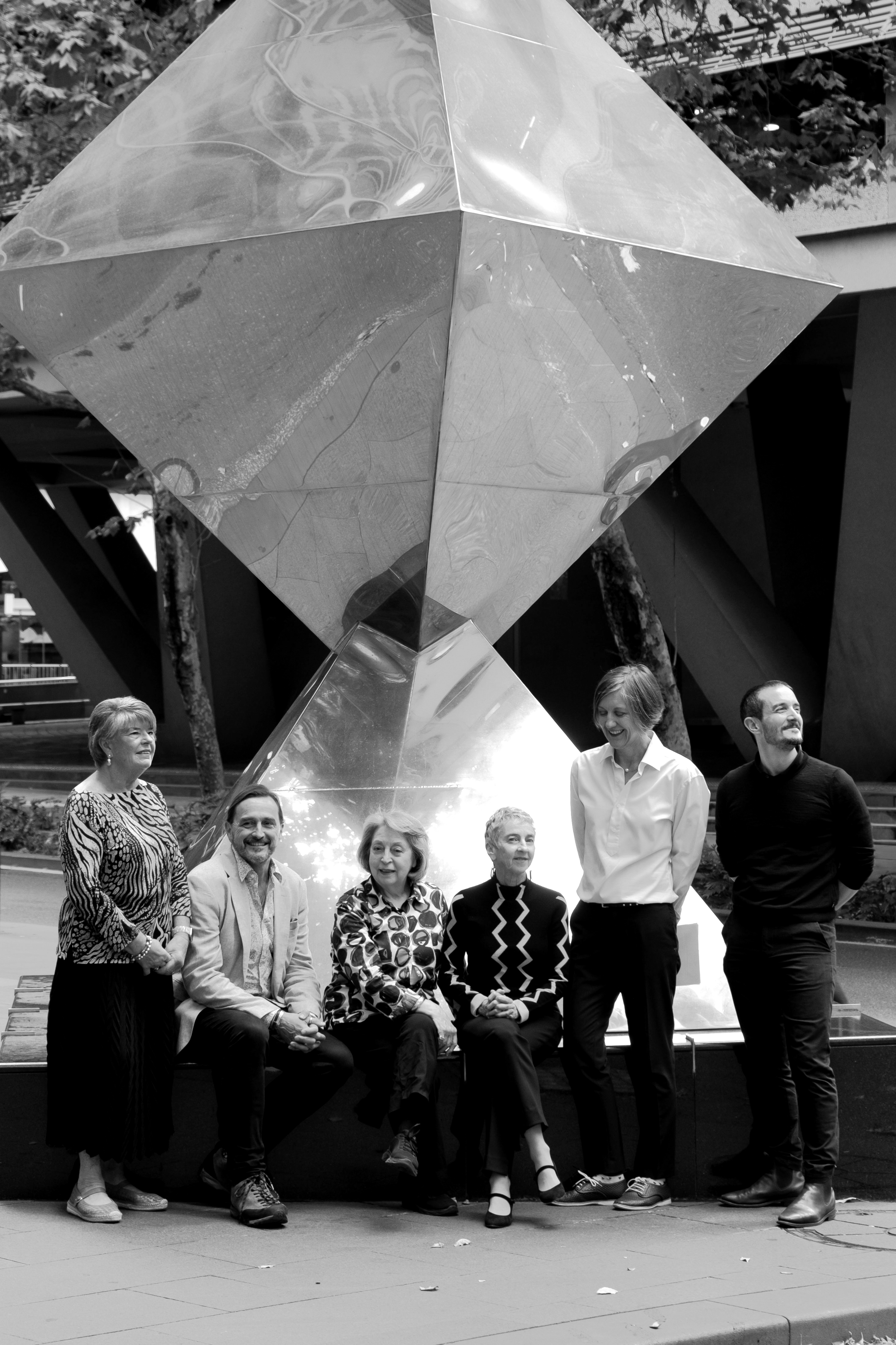 A group of people, six women and three men, pose in front of a large geometric reflective sculpture outdoors on a sunny day. The group includes a woman with short hair, a woman with longer hair, two women with patterned blouses, and three men, one with a beard and glasses. The sculpture is a shiny, multifaceted metal structure with reflections of the surroundings and people.