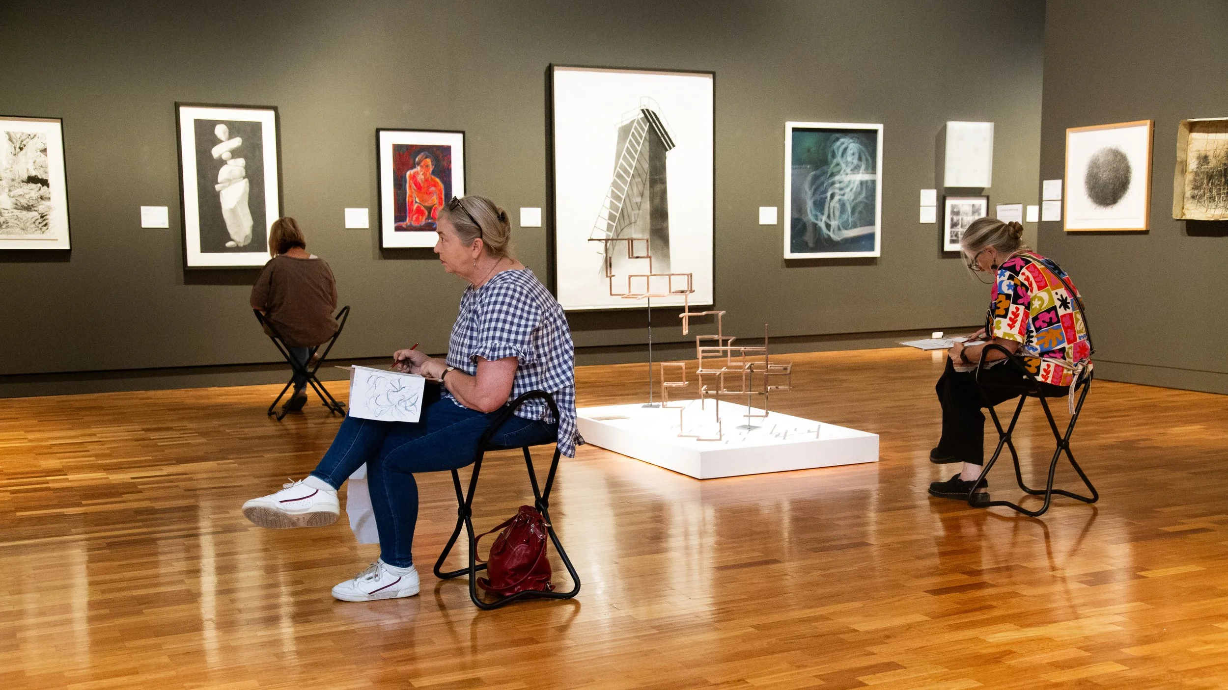 People sitting on chairs in an art gallery, looking at framed artwork on the wall and taking notes, with a sculpture in the center.