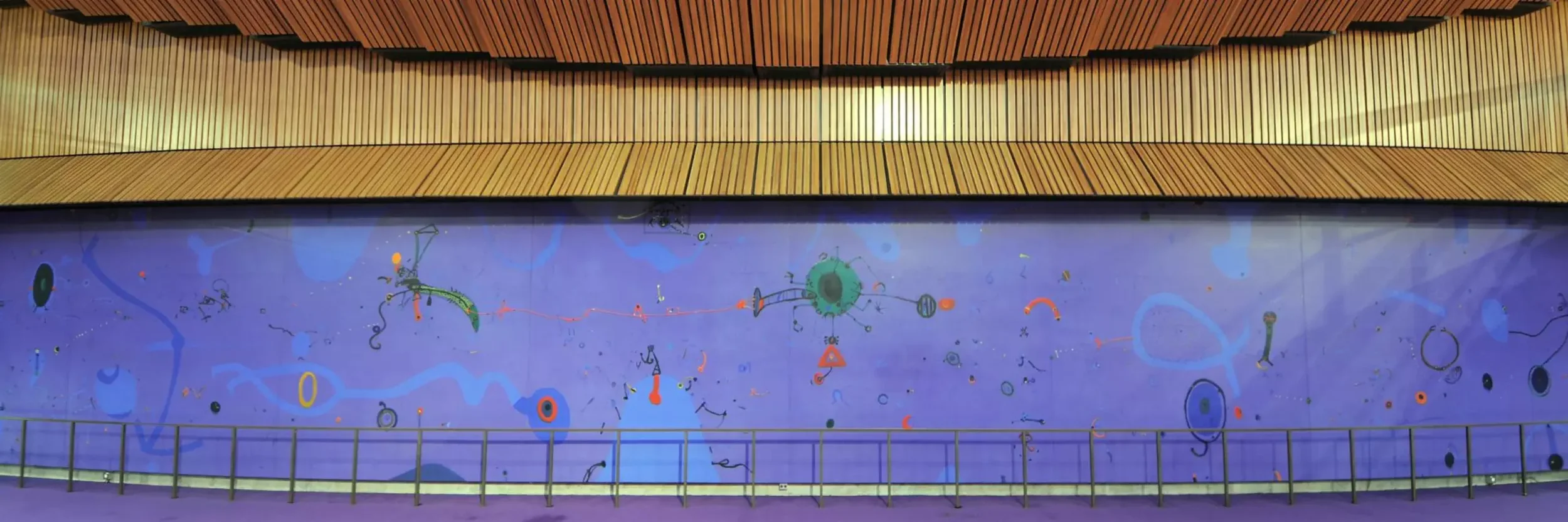 Colorful digital art mural with abstract shapes and patterns on a purple background, located inside a building with wooden panels on the ceiling.