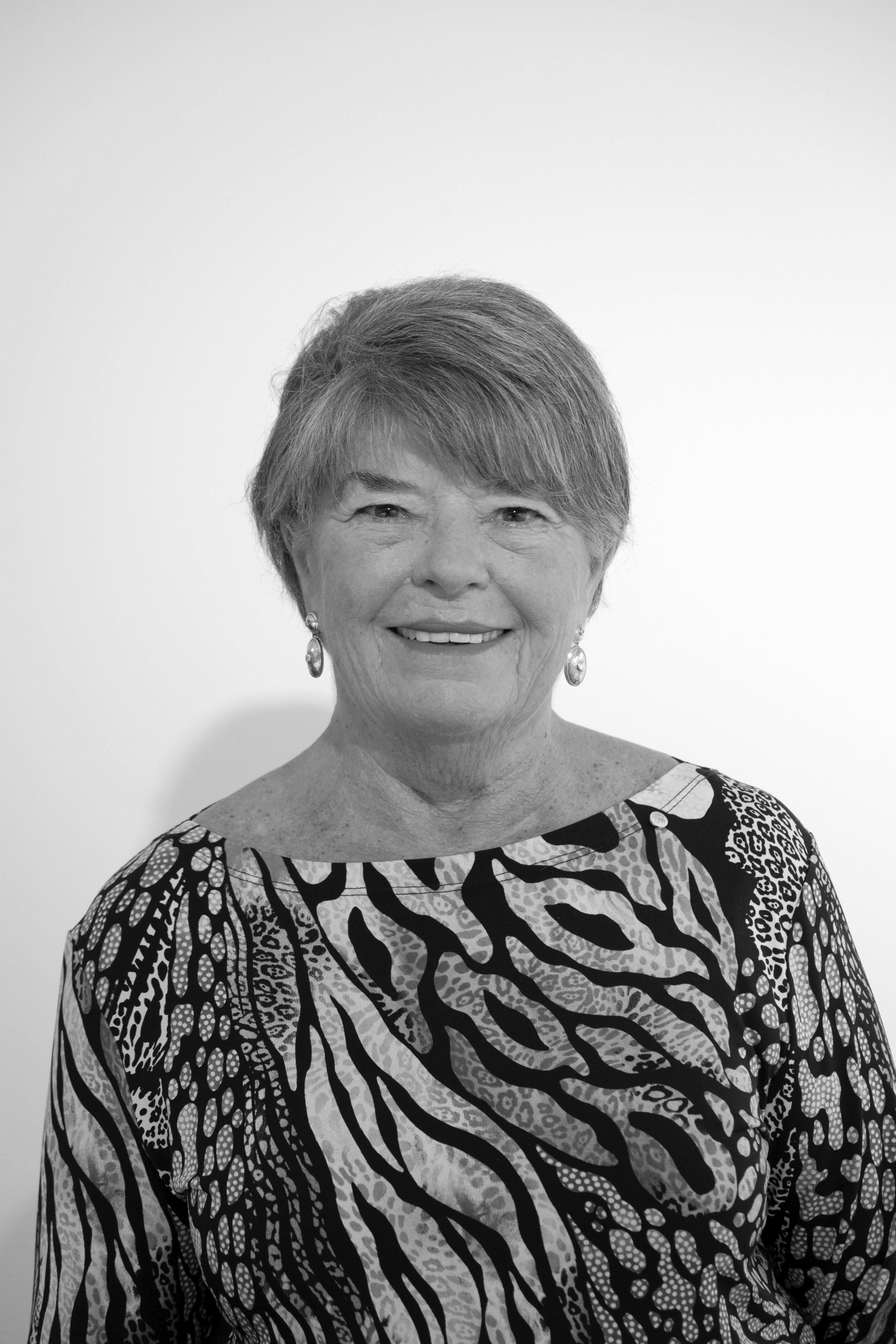 Black and white portrait of an elderly woman with short, layered hair, wearing earrings, and a patterned top with animal print and abstract designs, smiling against a plain light background.