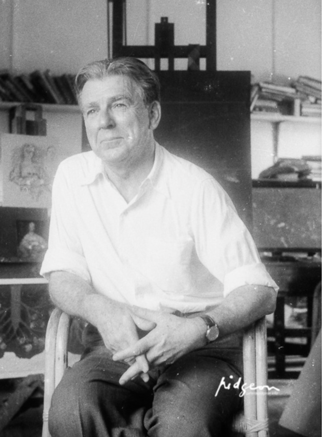 Black and white photograph of a middle-aged man with dark hair, sitting on a chair with one hand resting on his lap. The background features shelves with books and artwork, indicating an indoor setting.