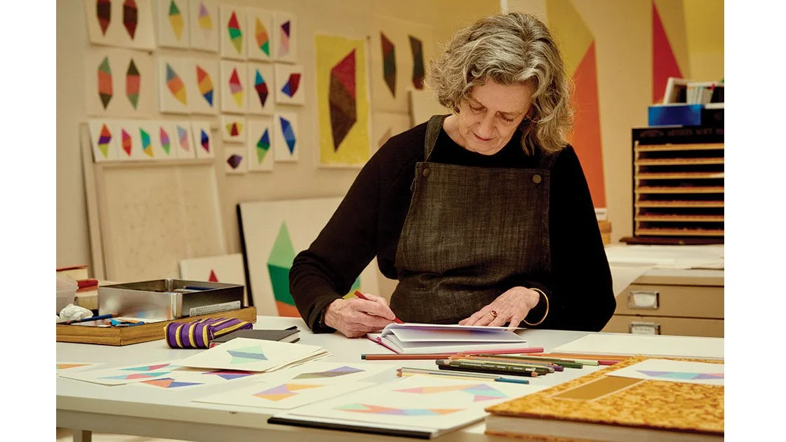An older woman with curly gray hair looking down and working on geometric art projects using colored pens in an art studio with colorful geometric artwork on the walls.