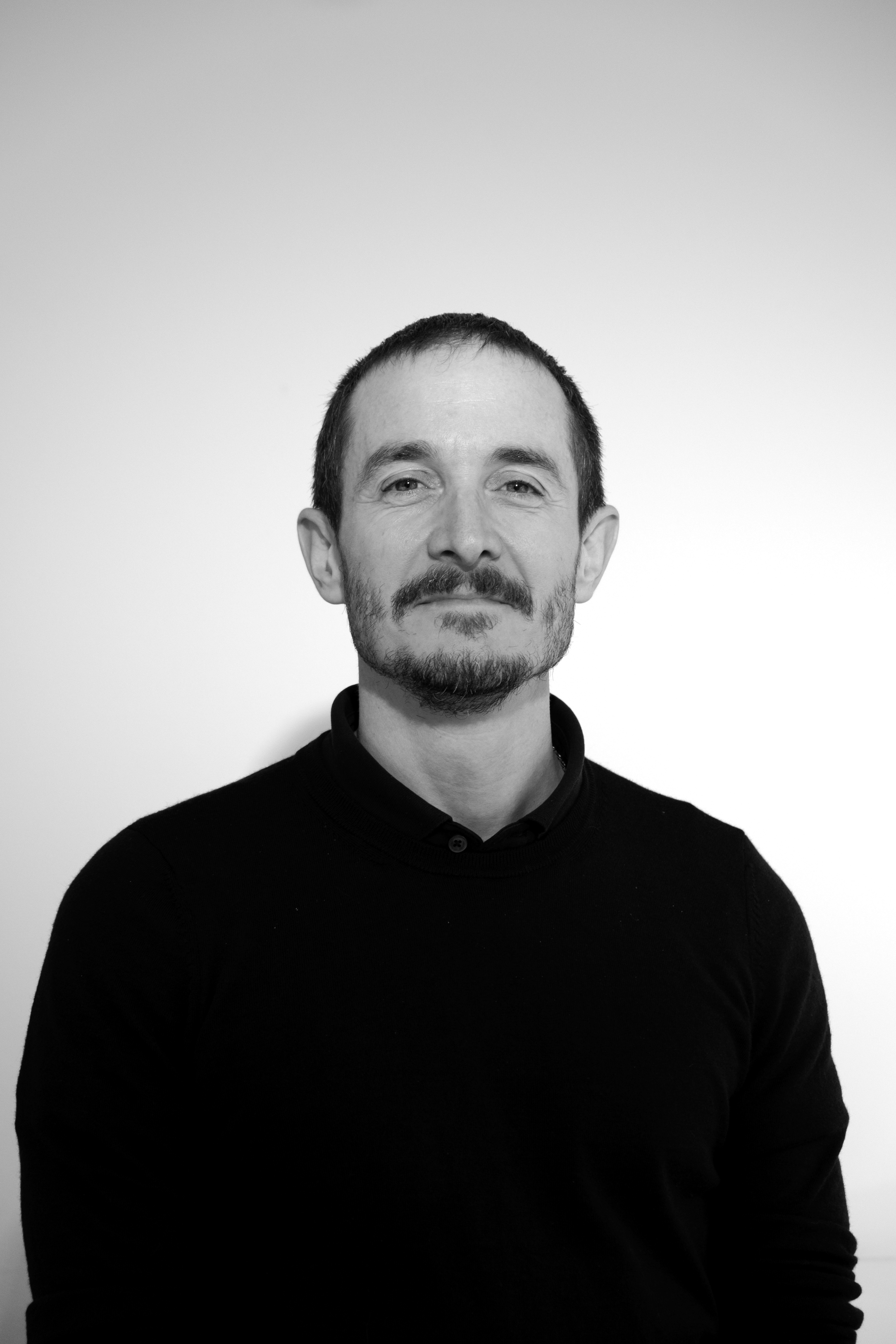 Black and white portrait of a man with short dark hair and facial hair, wearing a dark sweater, standing against a plain background.