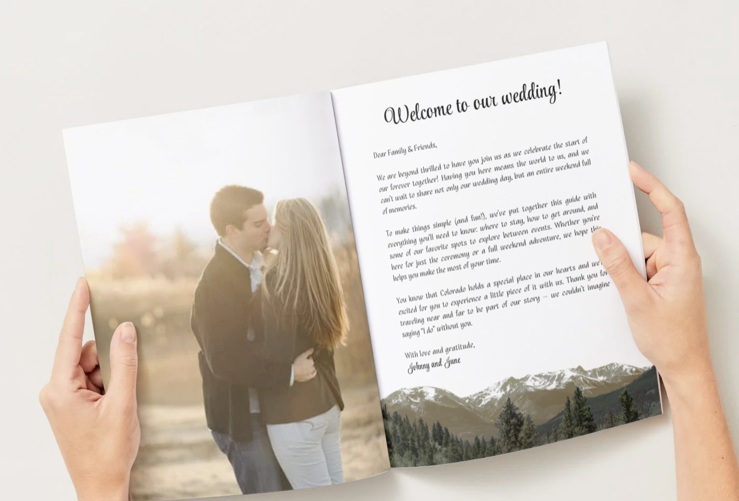 Hands holding an open wedding invitation with a photo of a kissing couple on the left page and a scenic mountain landscape at the bottom of the right page.