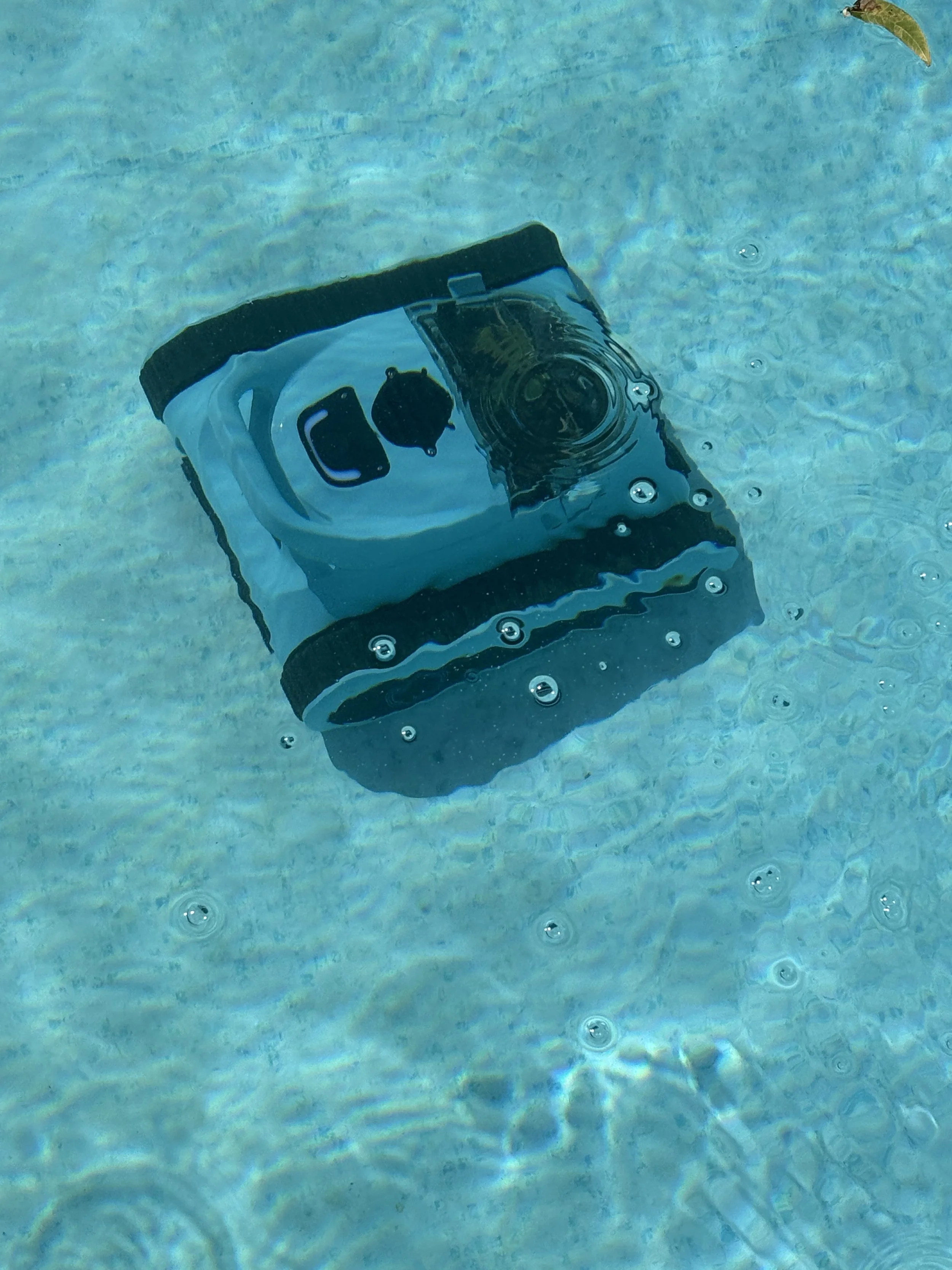 A waterproof action camera in a protective case submerged in clear water, creating ripples and bubbles around it.