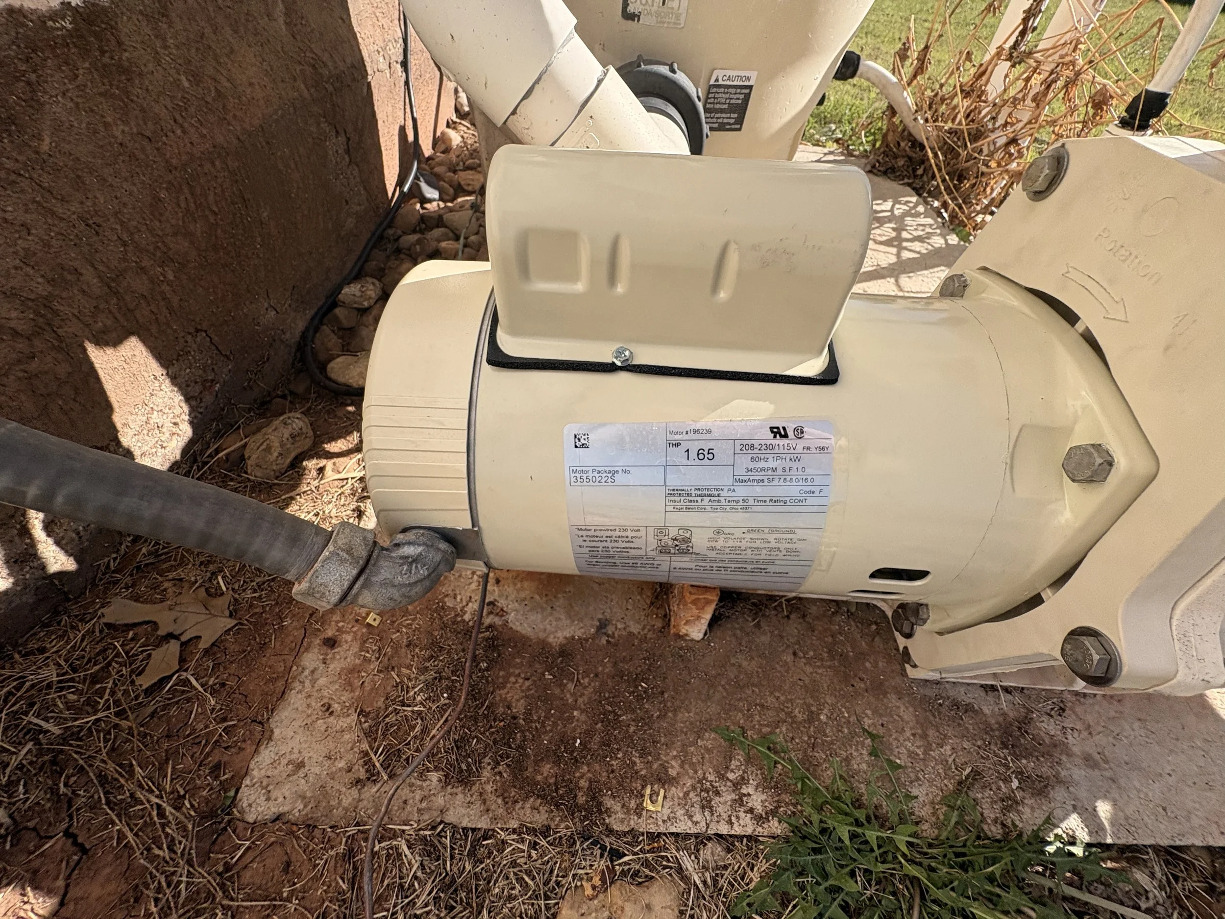 Mechanical equipment placed on the ground, connected via cables and pipes, with soil and rocks surrounding it and some plants nearby, possibly an outdoor utility or HVAC unit.
