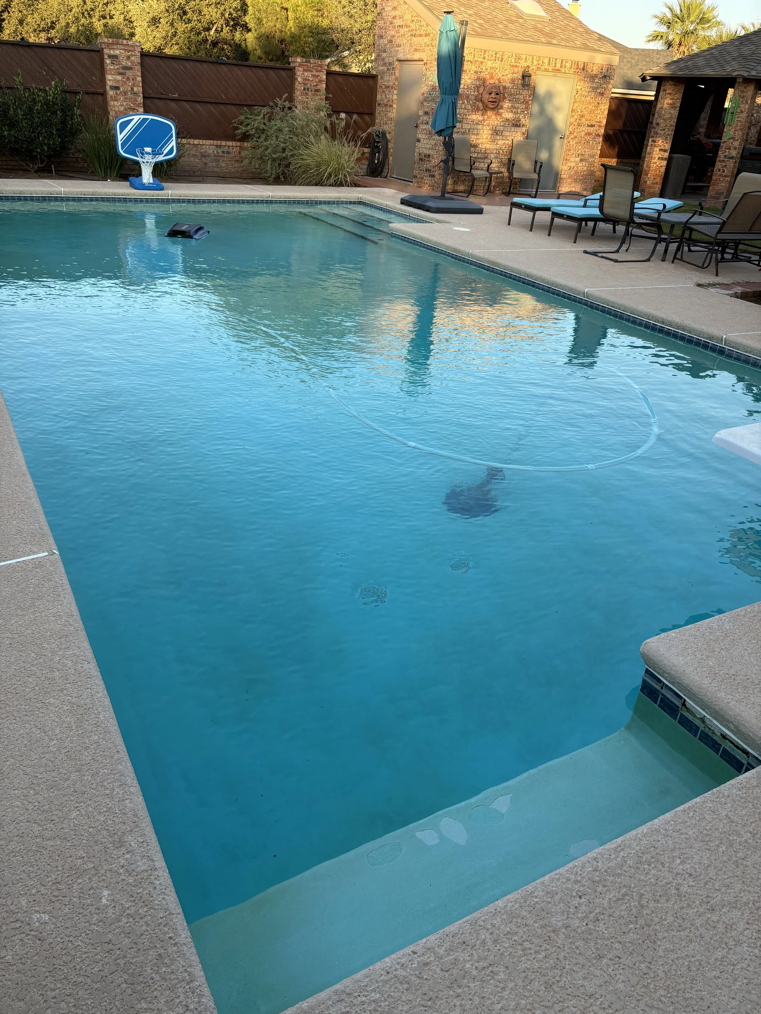 Backyard swimming pool with clear blue water, surrounded by concrete deck, poolside chairs, a divided blue basketball hoop, a small shed, a fence, and some plants. The setting appears to be during late afternoon or early evening.