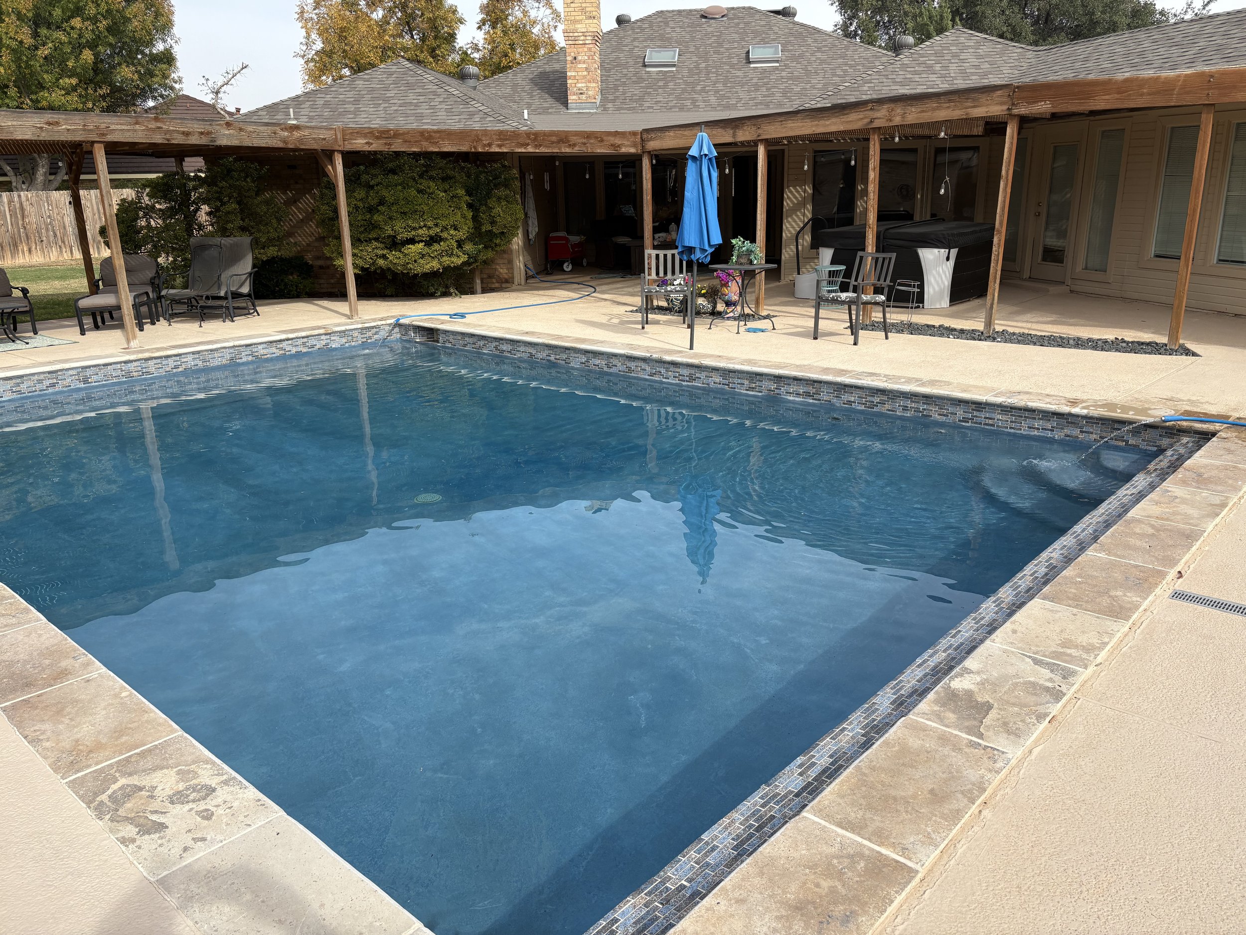 Backyard swimming pool with clear blue water, surrounded by beige and tan paving stones. The backyard has a wooden privacy fence, some outdoor chairs, a table with an umbrella, potted plants, and a covered hot tub. A house with a shingled roof and sliding glass doors is visible in the background.