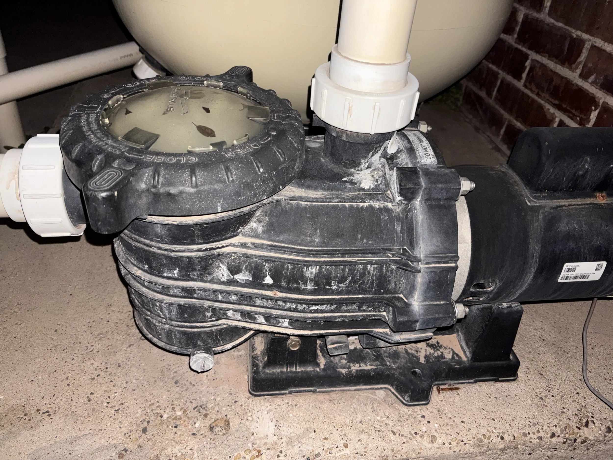 Close-up of an indoor pool pump with a dirty black casing, white pipes, and a grime-covered surface, with a brick wall in the background.