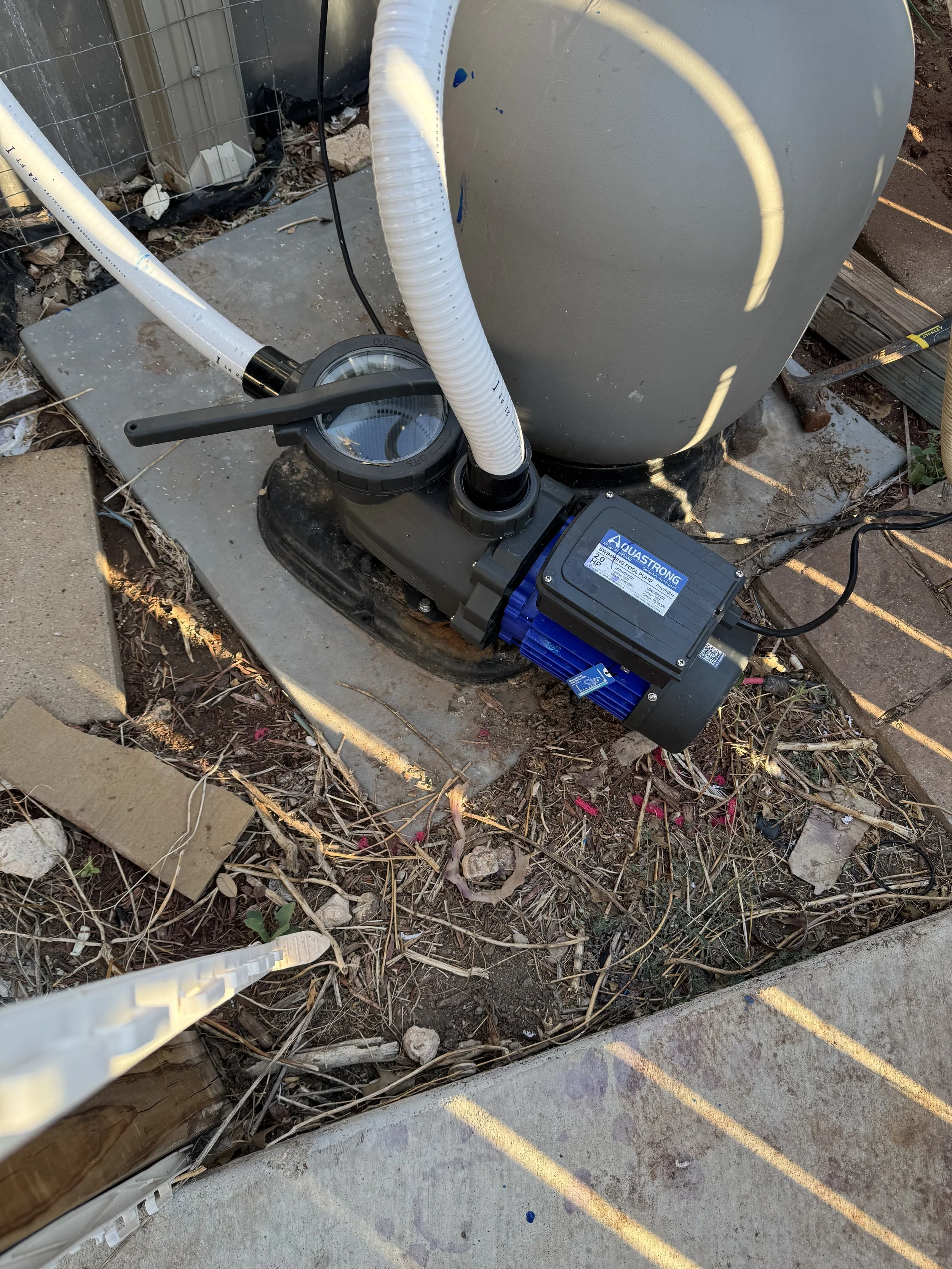 Close-up of a pool pump installed outdoors, with pipes and wires connected, surrounded by dirt, debris, and a concrete surface.