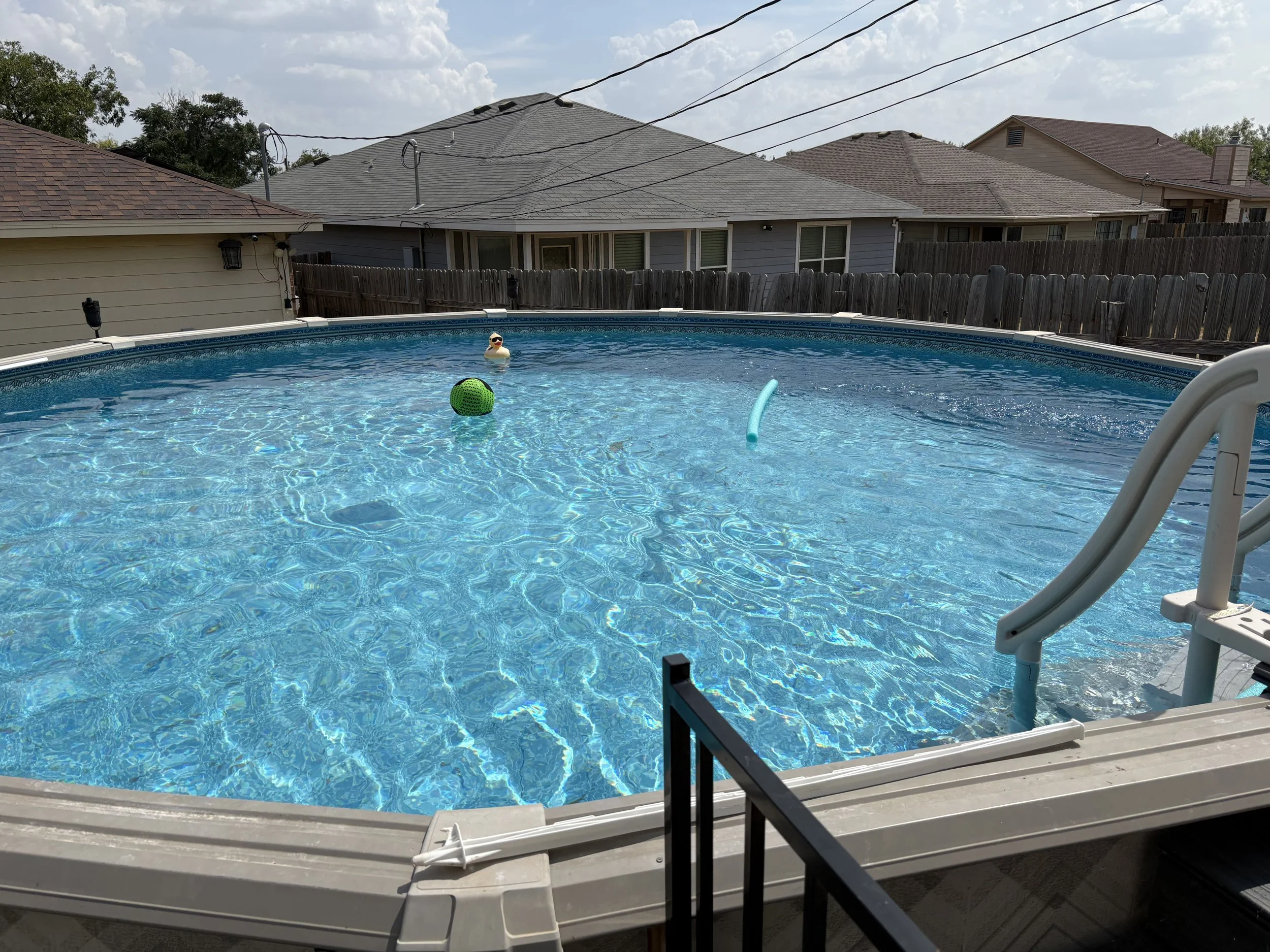 A backyard swimming pool with a lounge chair in the foreground and a metal railing. The pool water is blue and has two floating toys, one shaped like a duck and the other like a ball. Surrounding the pool is a wooden fence, with neighboring houses and power lines visible in the background under a partly cloudy sky.